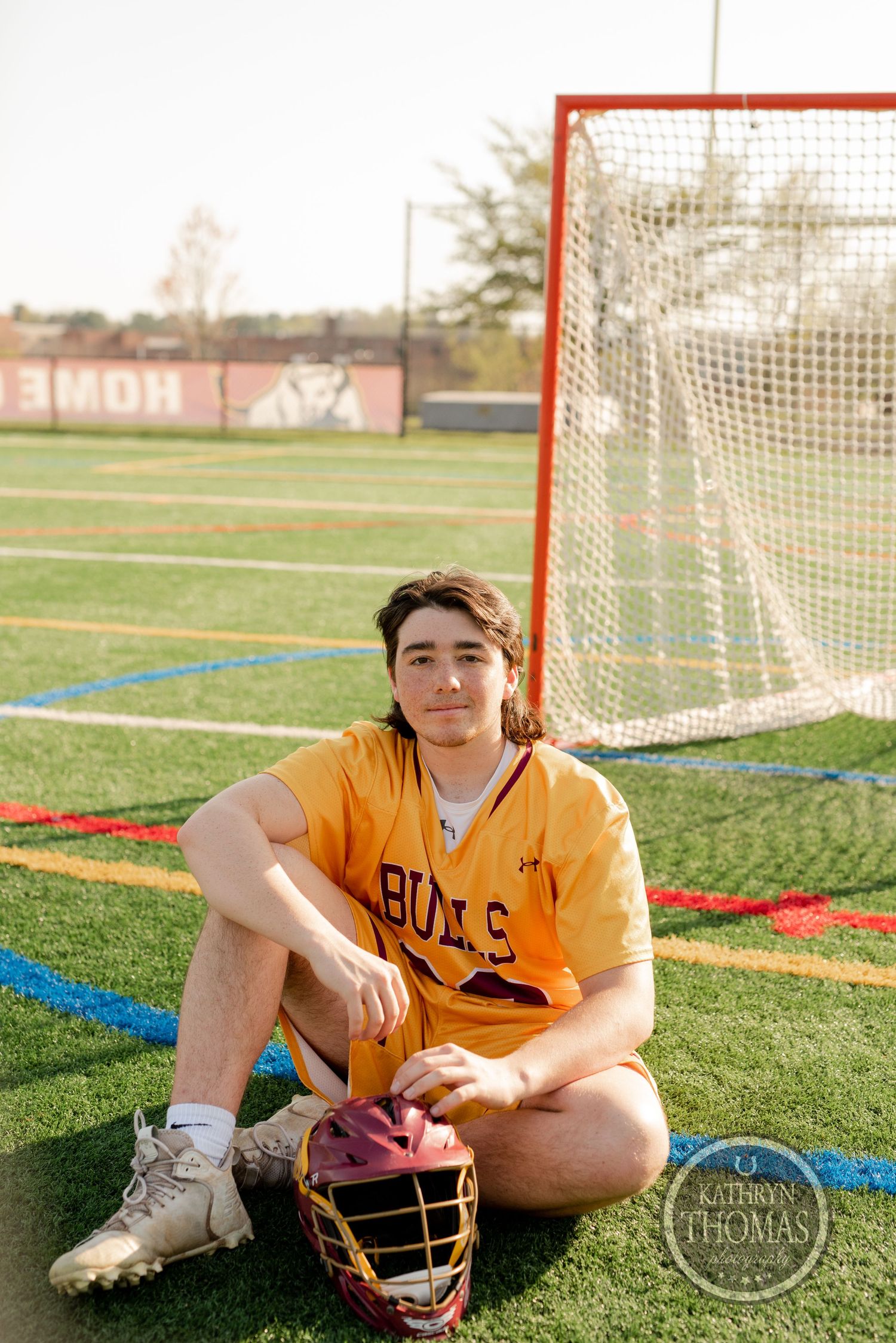 Athlete in yellow Bulls jersey sitting casually with lacrosse helmet and stick by goal.