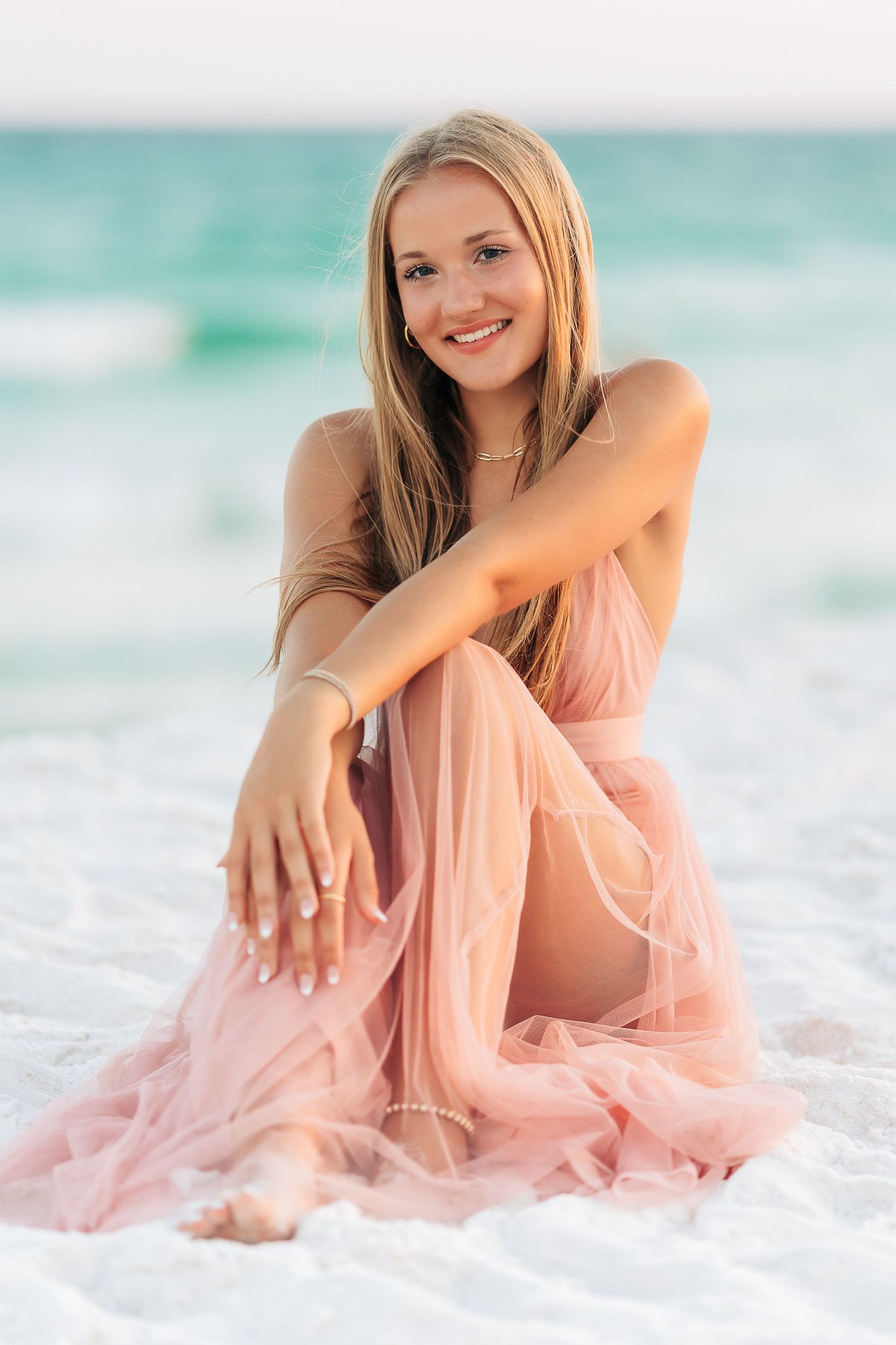 Woman in flowing pink dress poses gracefully at sunset on beach as waves lap at shore in dreamy seaside photo session.Navarre Beach