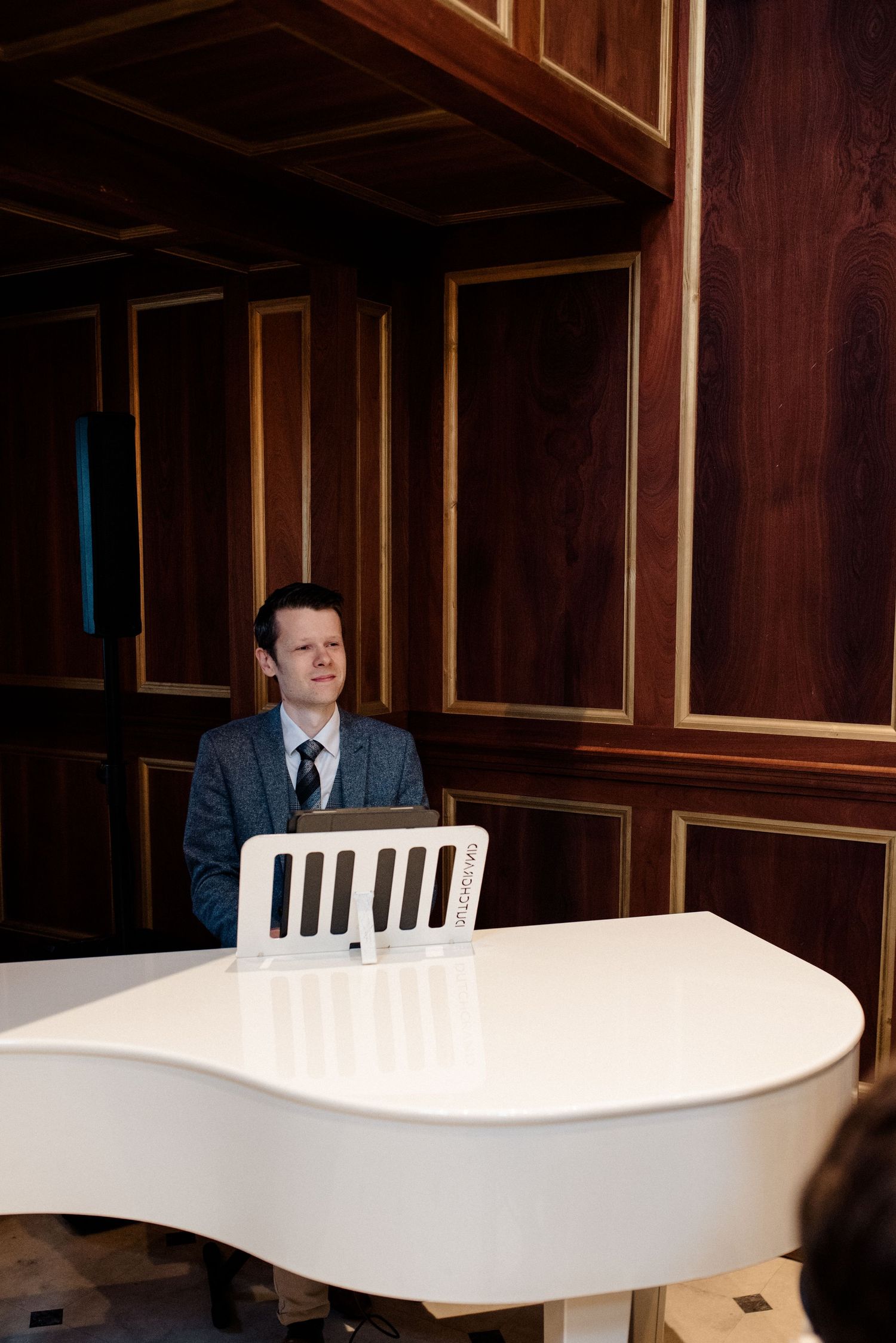 Pianist in a grey suit sitting at a white grand piano in an elegant wood-paneled room.