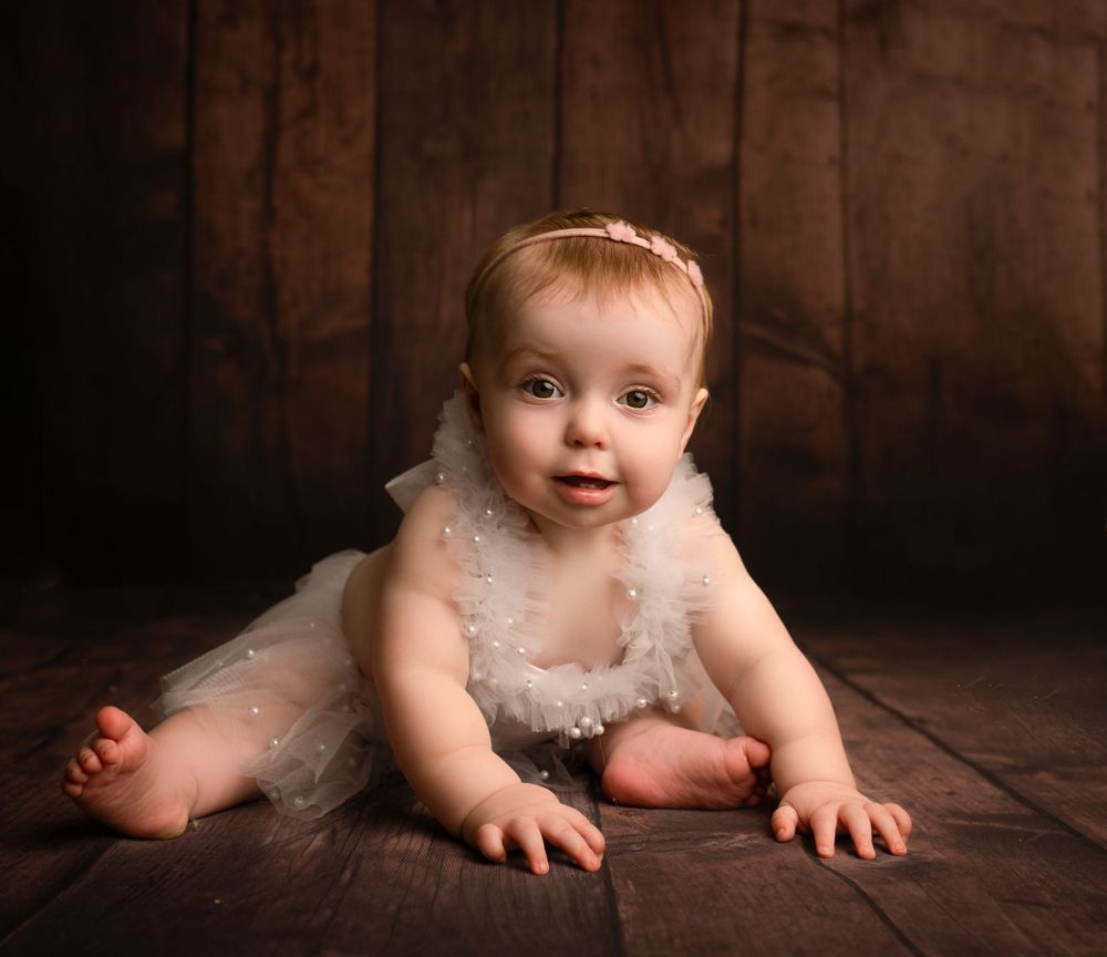 baby girl in white dress sat up smiling on wooden backdrop photographed by newborn photographer swansea