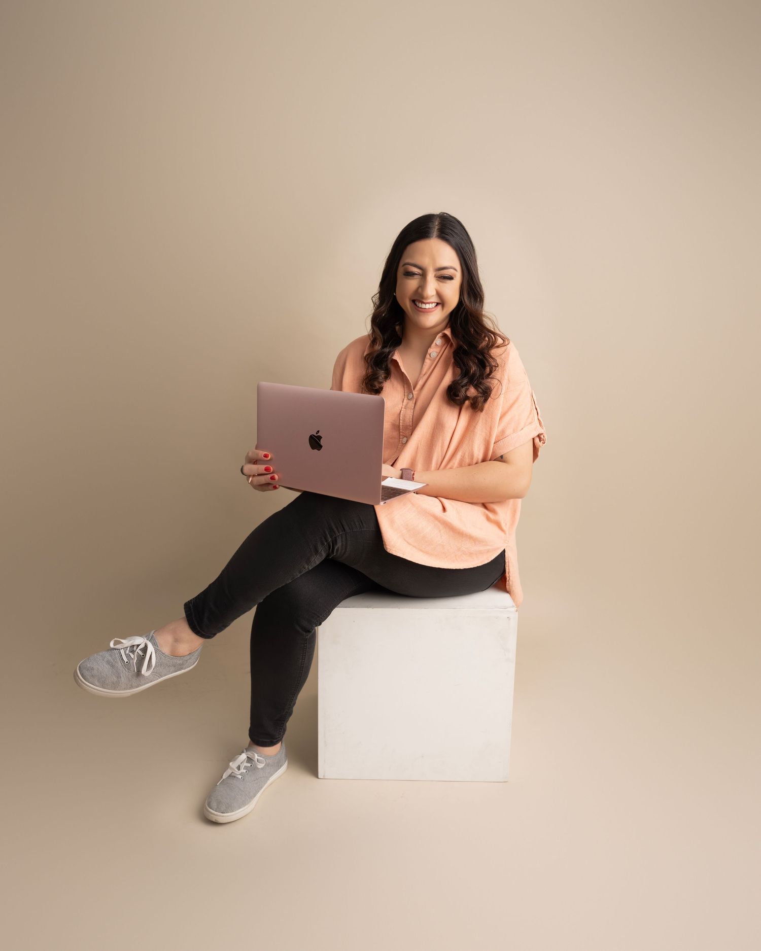 Casual business photo with person in peach sweater using laptop while seated on white cube.