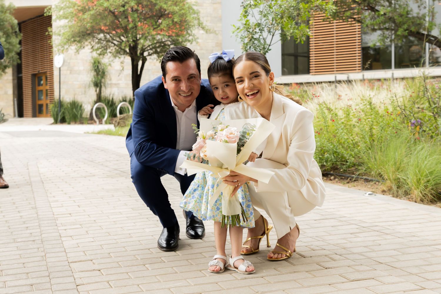Smiling couple crouches down with young girl holding flowers on sunny outdoor pathway.
