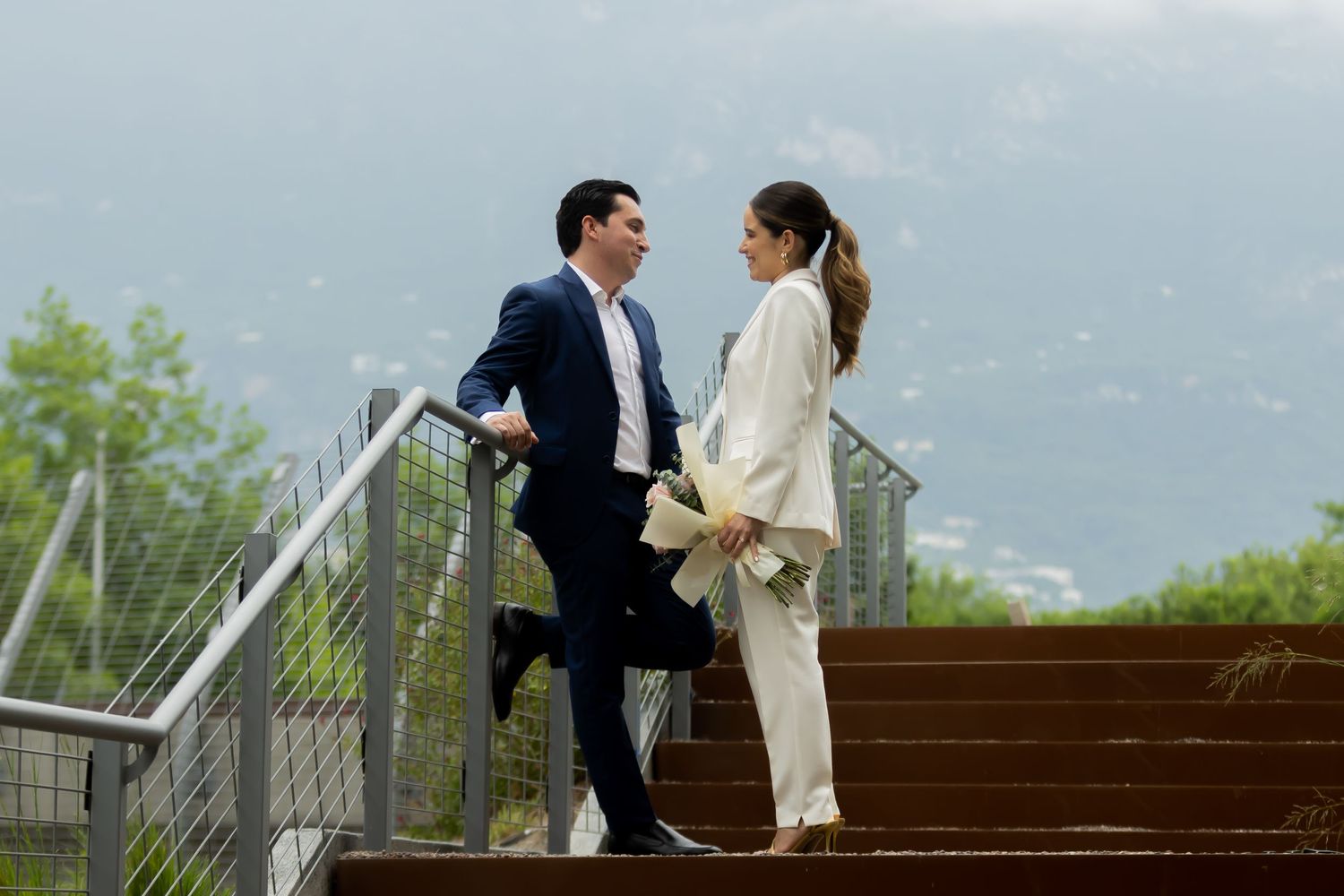 Bride and groom in white suit exchanging smiles on wooden steps with mountain view backdrop.
