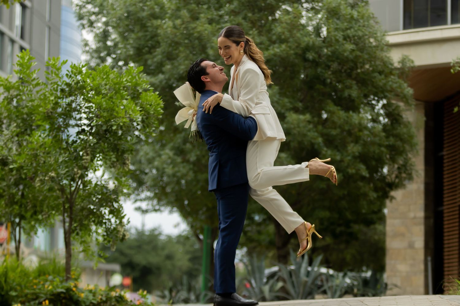 Groom lifting bride in white suit outdoors during joyful wedding photo session.