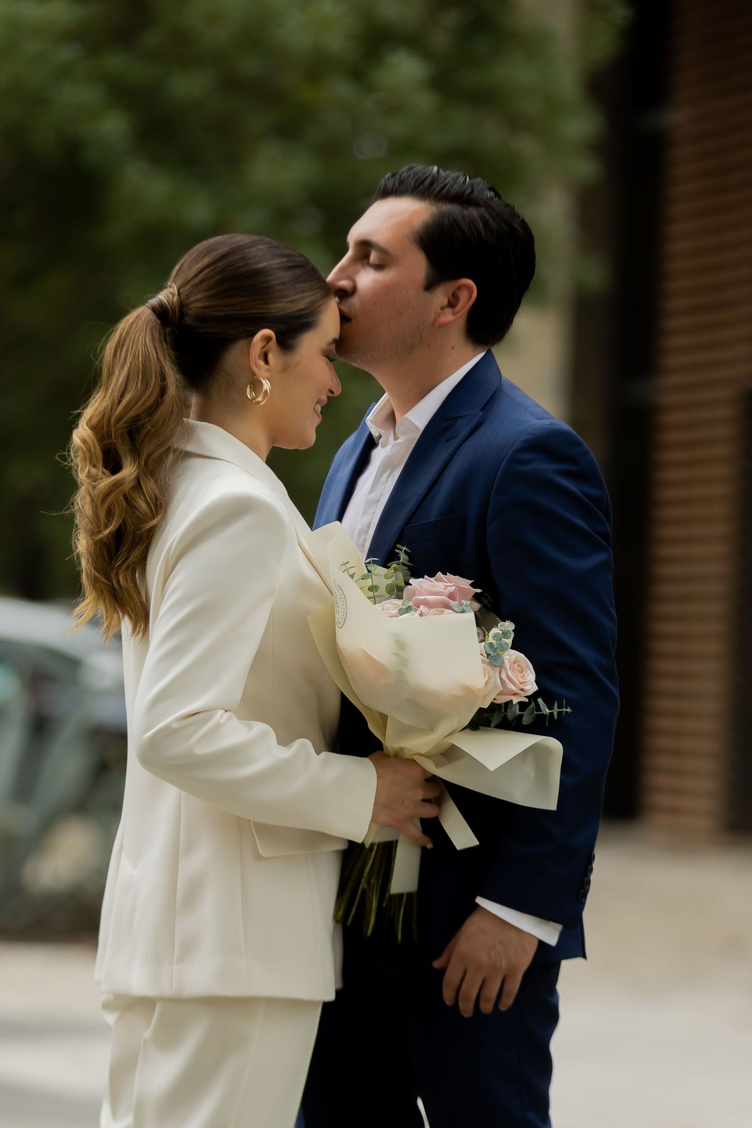 Romantic couple sharing a kiss while holding a pink rose bouquet on a city street.