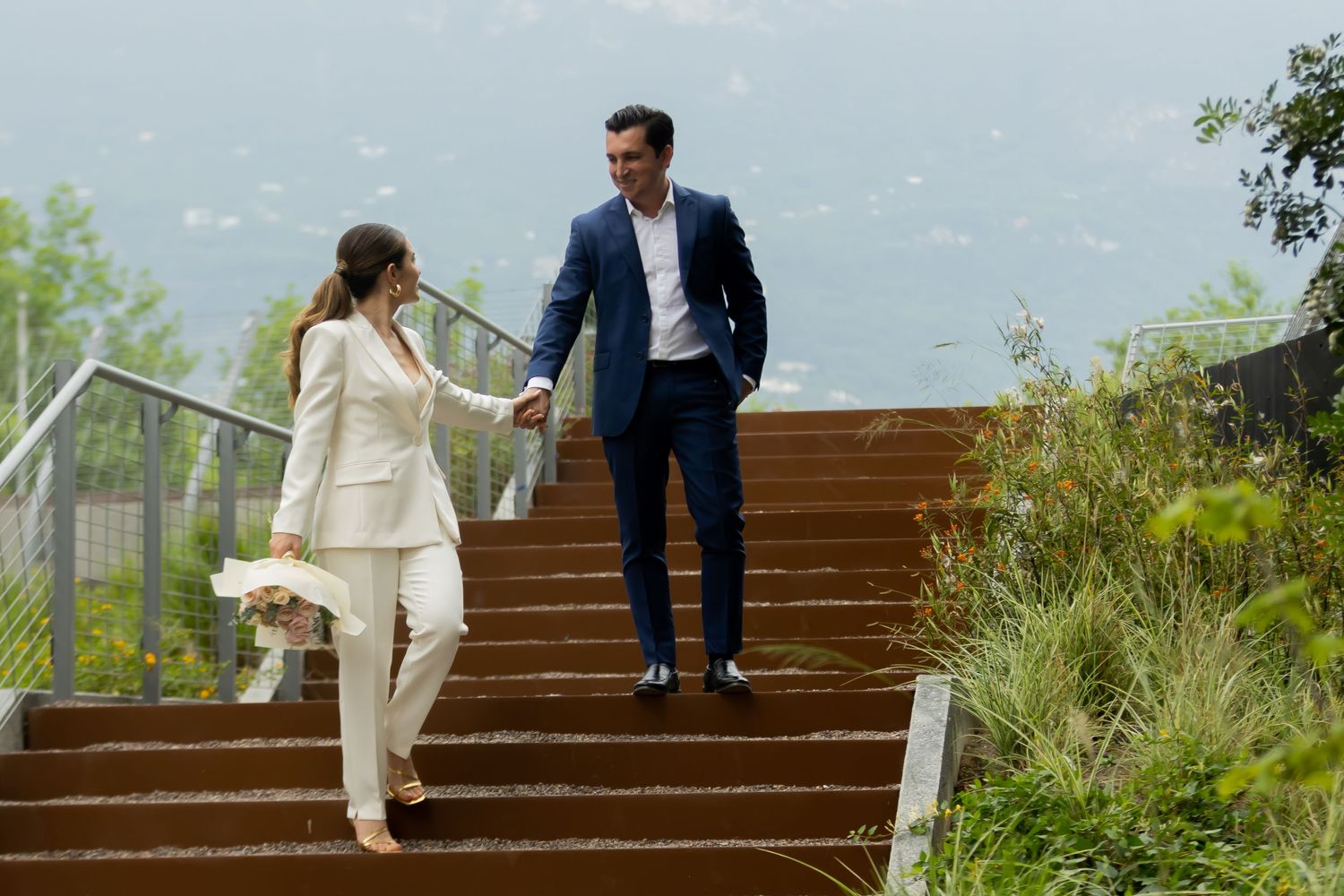 Elegant couple in formal attire holds hands while climbing wooden stairs with scenic view.