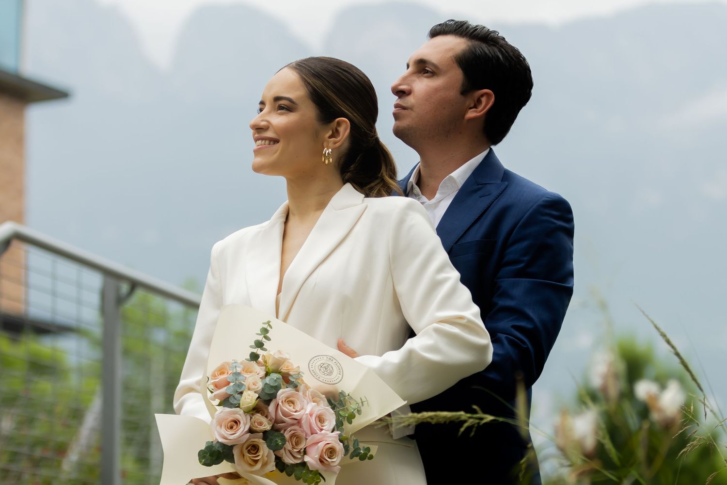 Elegant couple in white suit and navy blazer posing with bouquet at scenic mountain wedding.