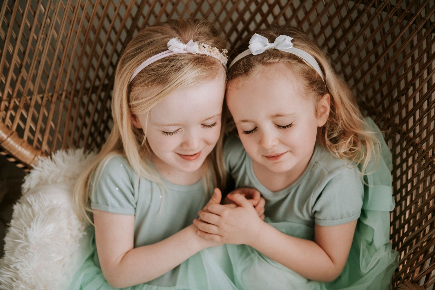 Two young girls in matching mint green dresses share a sweet moment together on a wicker chair.