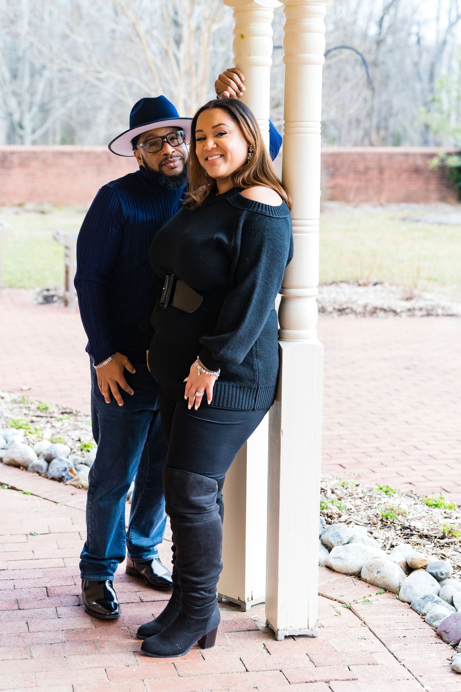 A couple in matching dark sweaters pose together by a white pillar on a brick patio.