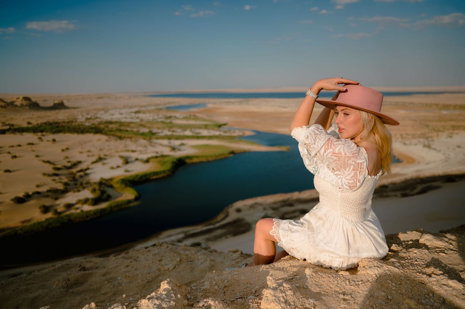 Woman in white dress sits on cliff edge overlooking Lake Powell on a sunny day.