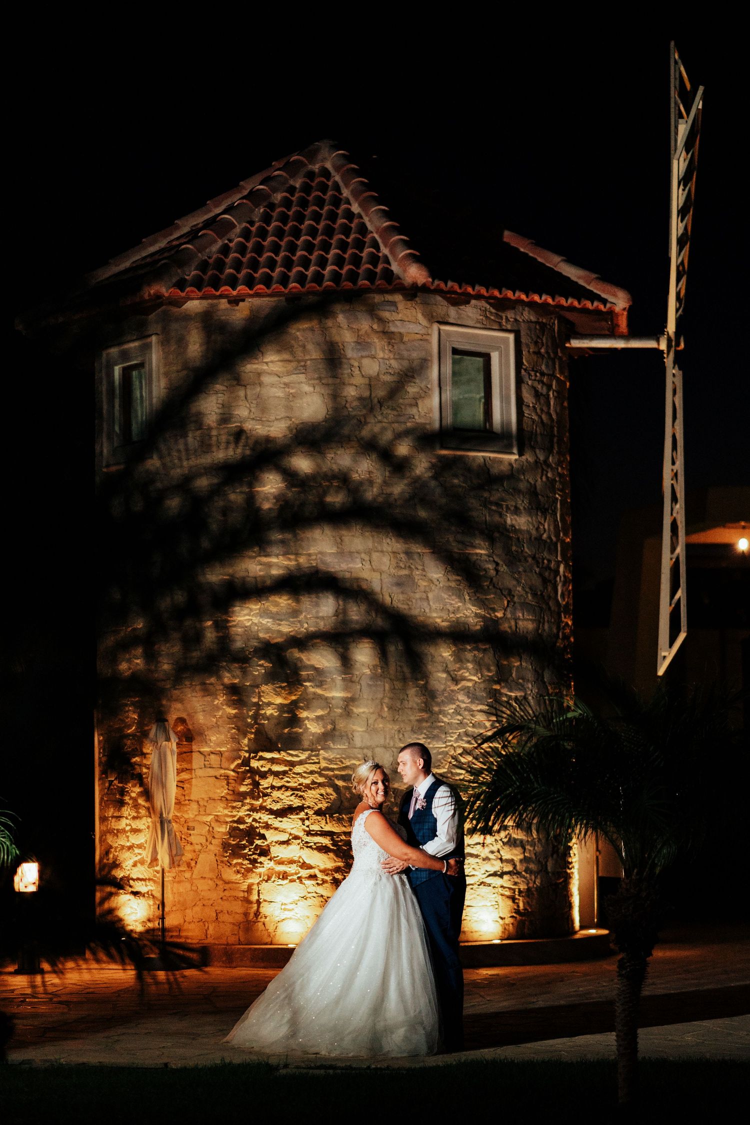 A wedding couple poses at night against a dramatically lit stone tower with a tiled roof.