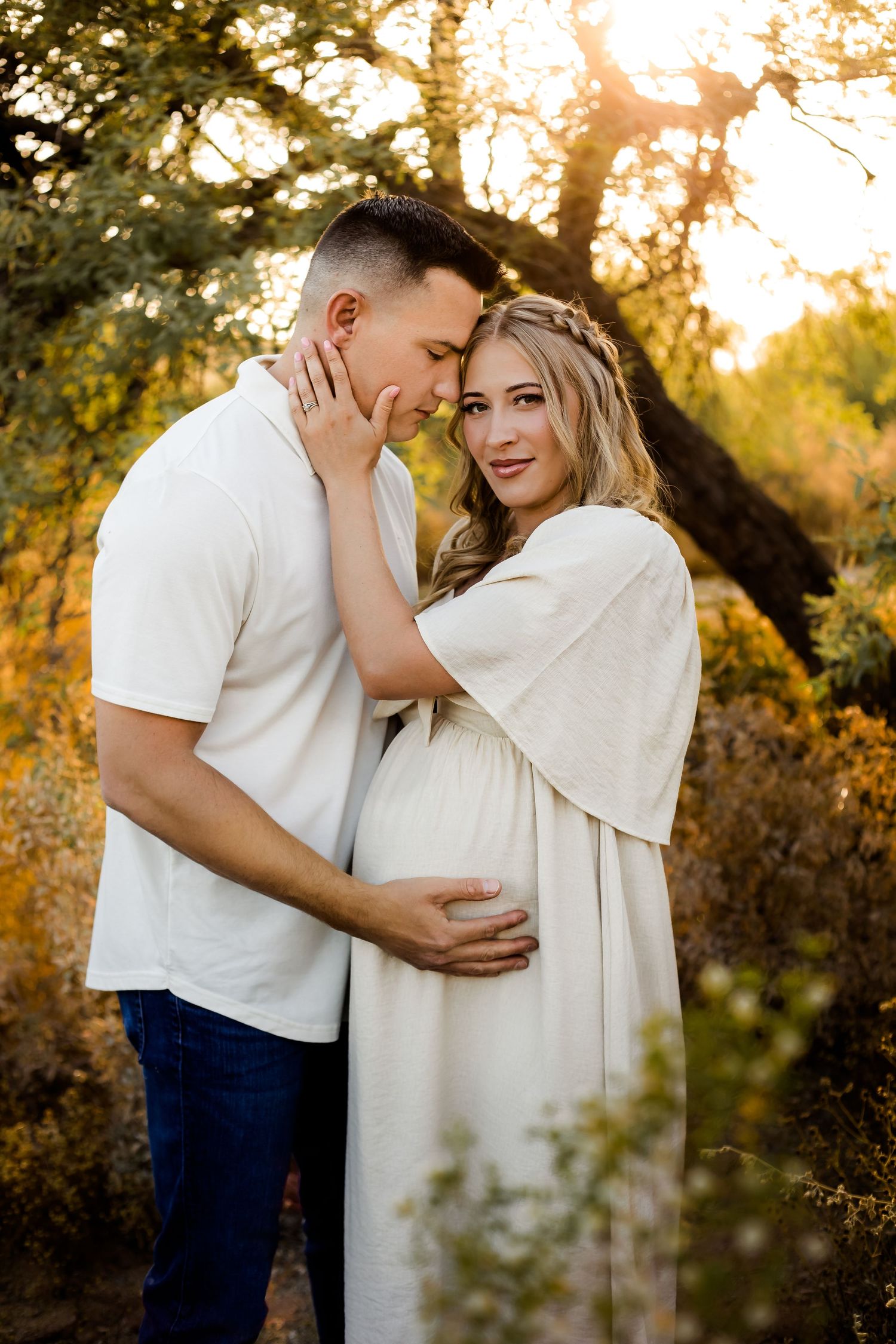 Stunning maternity mom stares at the camera while her husband snuggles her and her baby belly in the golden glow of the Arizona desert.