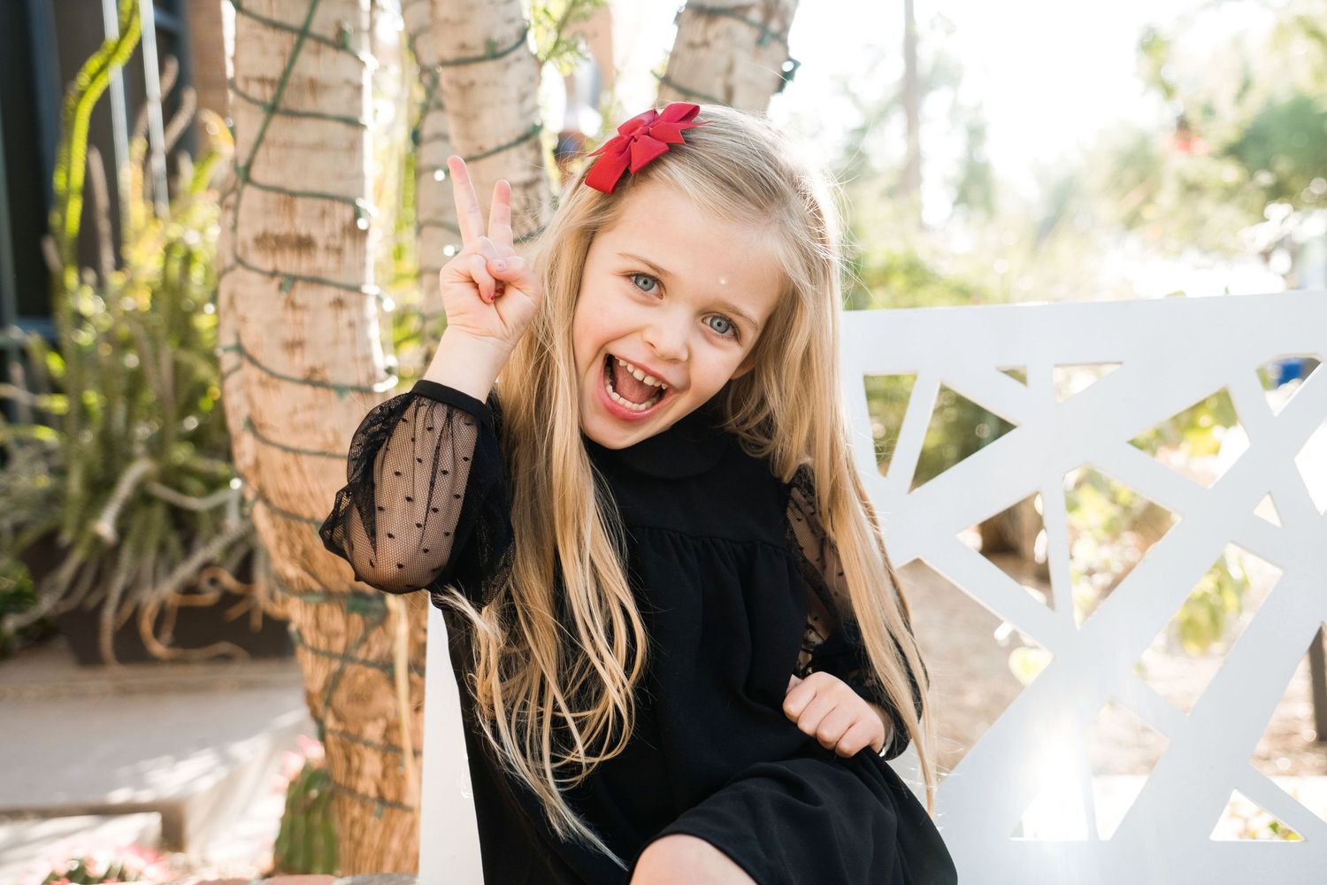 Young child in black dress poses playfully against white fence and foliage.