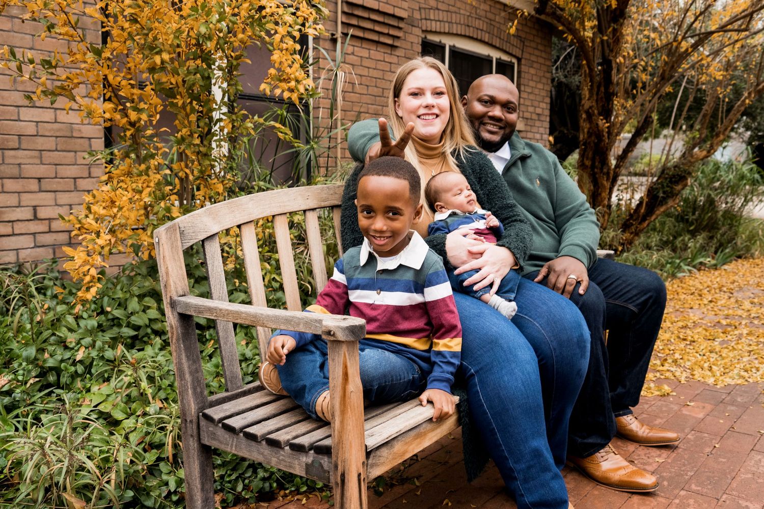 Family sitting together on a wooden bench surrounded by fall foliage on a brick patio.
