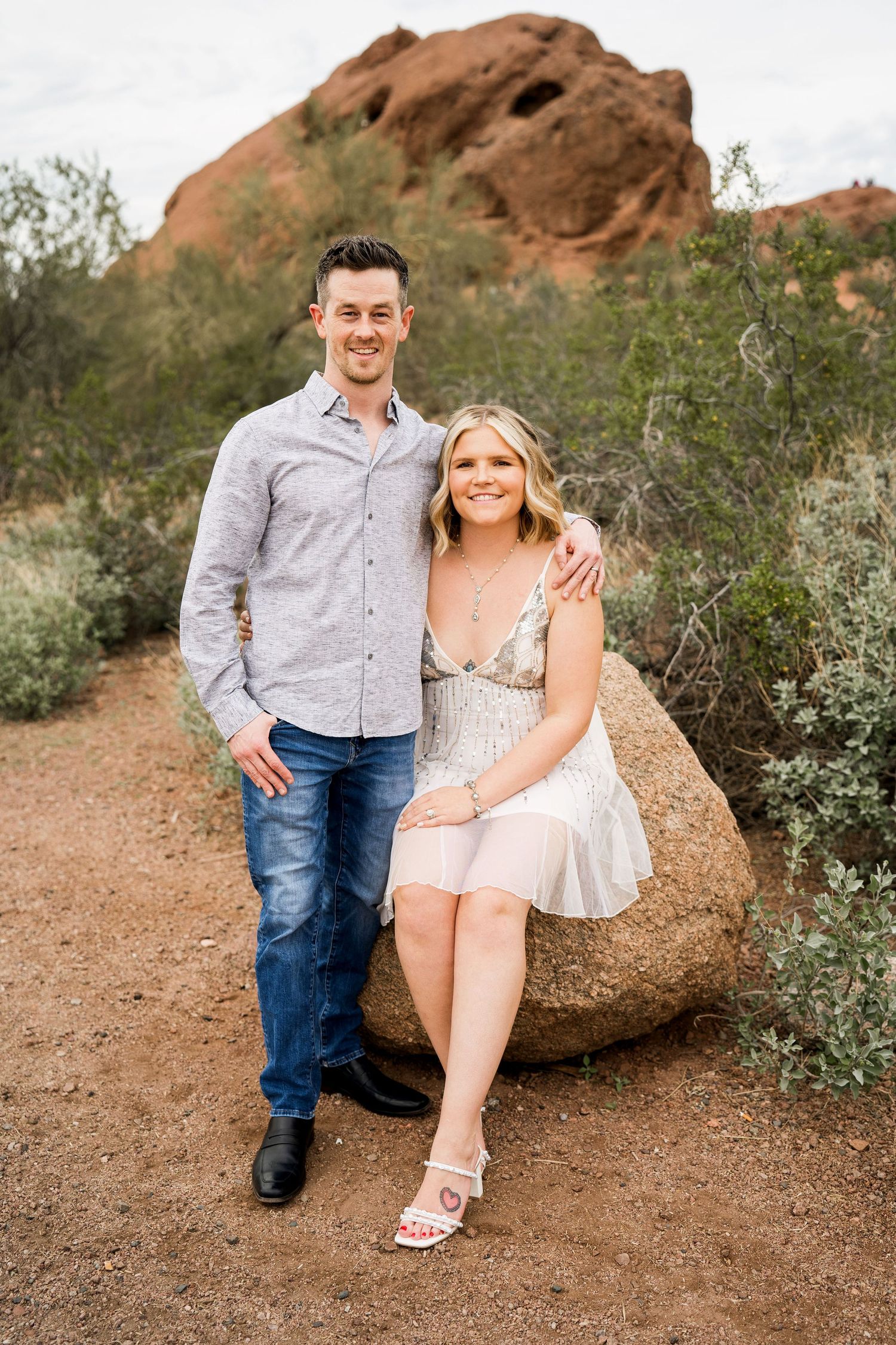 Couple sitting on boulder with red rock formation backdrop in desert landscape.