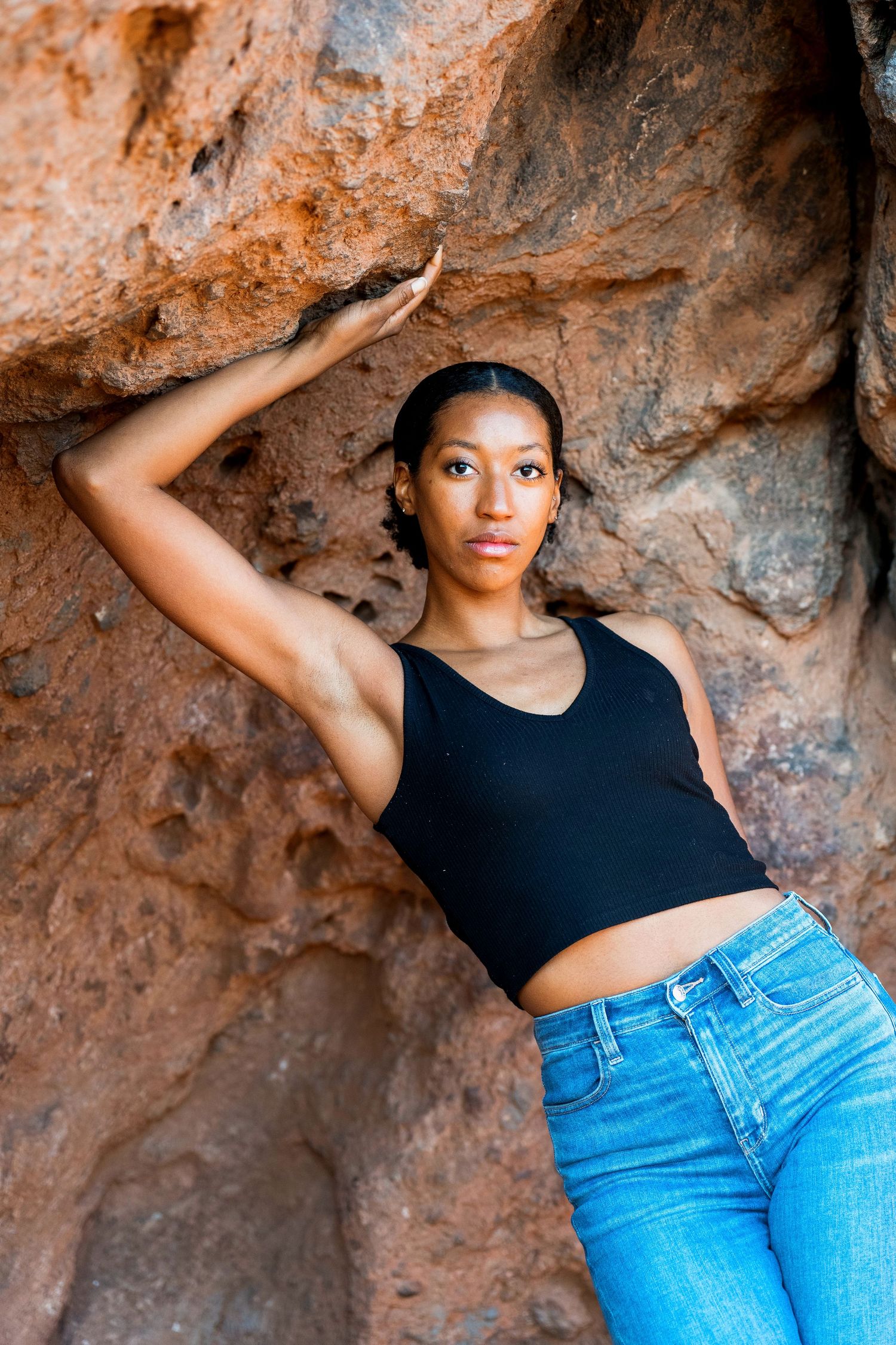 Athletic woman in black top and jeans poses against red rock formation.