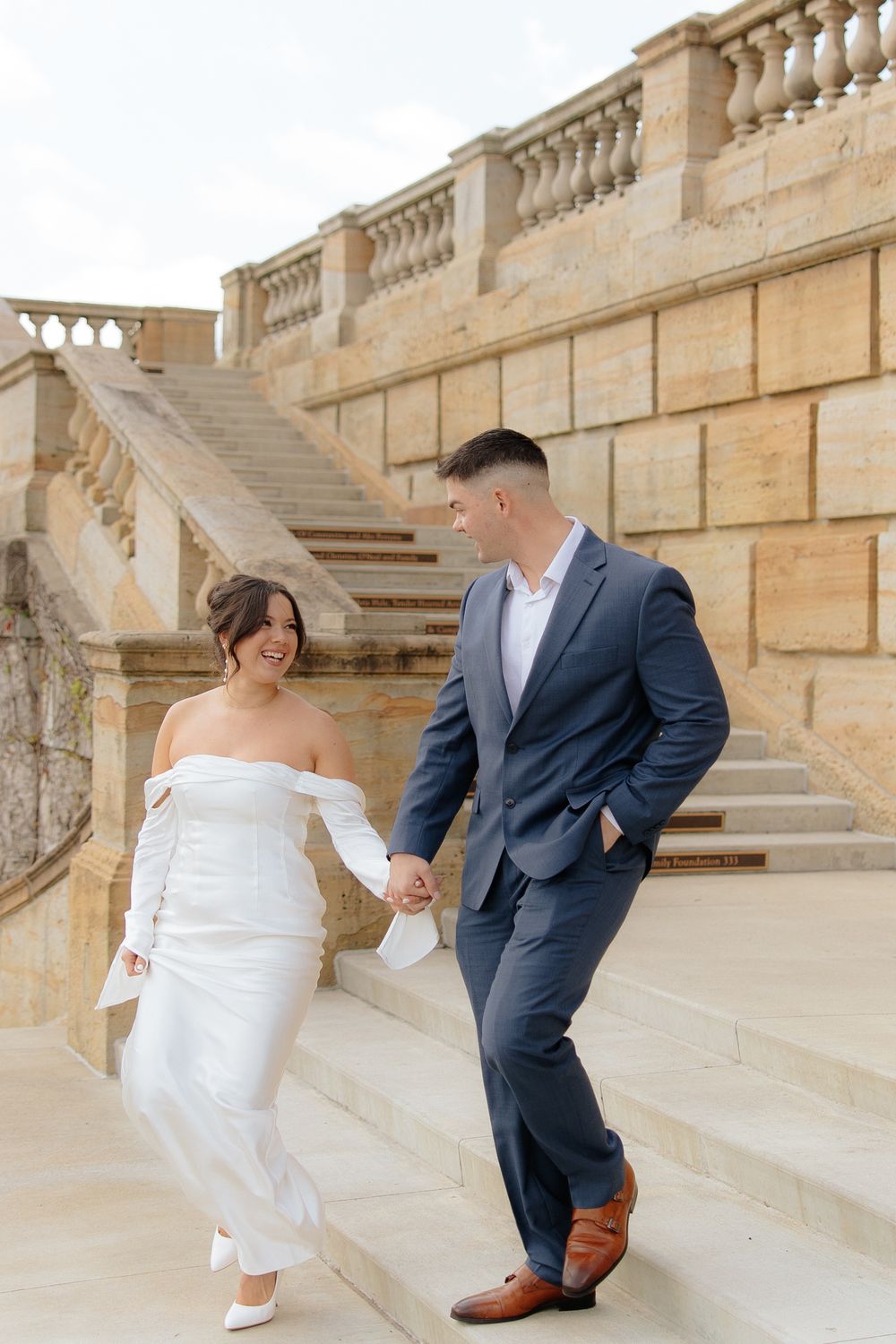 A newlywed couple in formal wedding attire pose together on elegant stone stairs with ornate balustrades.