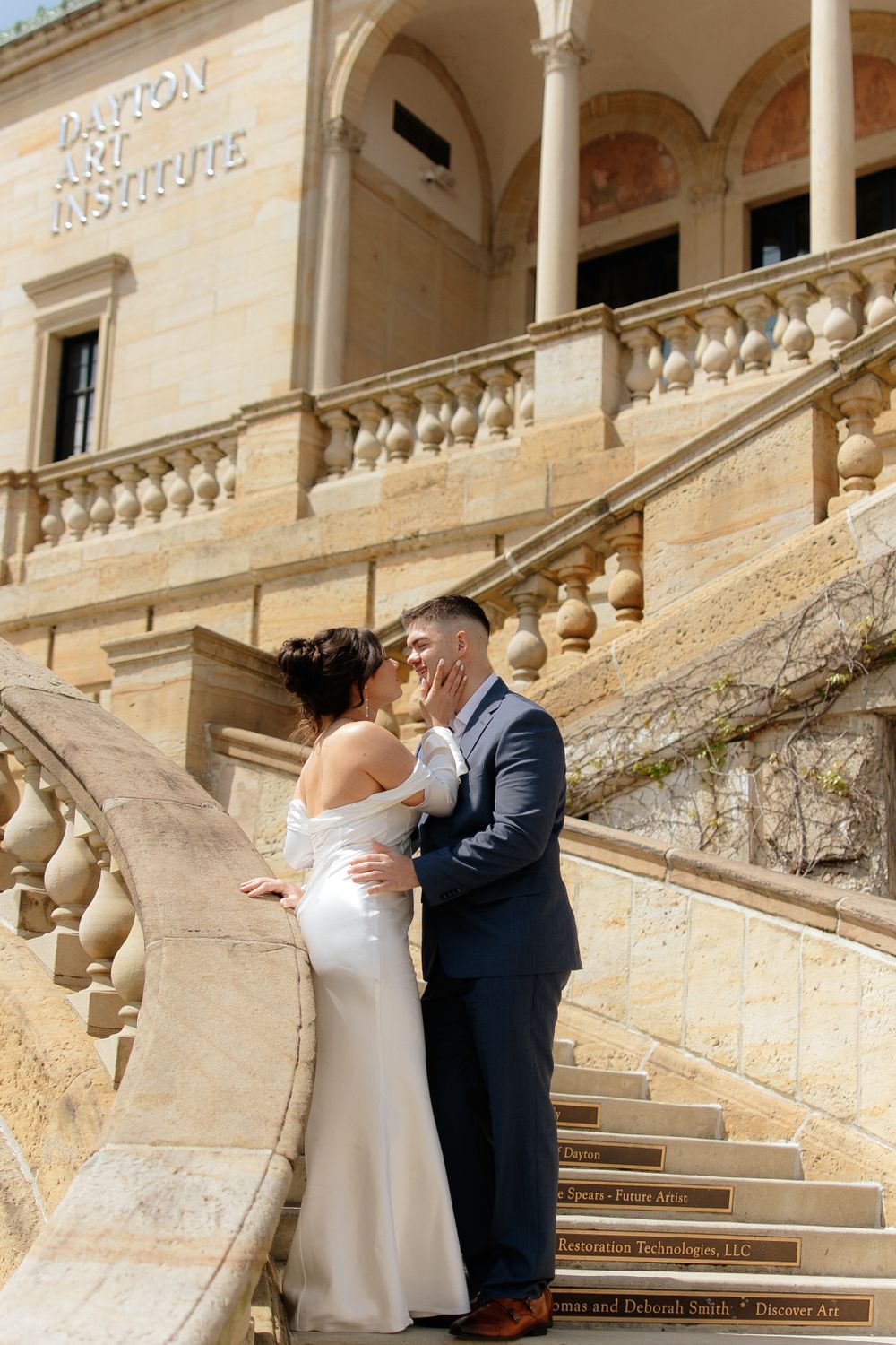 A romantic wedding couple embraces on an elegant stone staircase with classical architecture and balustrades.