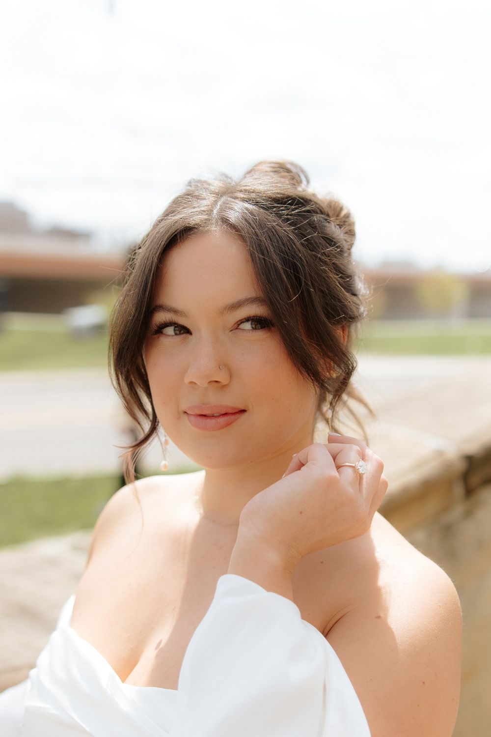 Portrait of someone in a white off-shoulder dress with soft makeup and an upswept hairstyle in natural lighting outdoors.