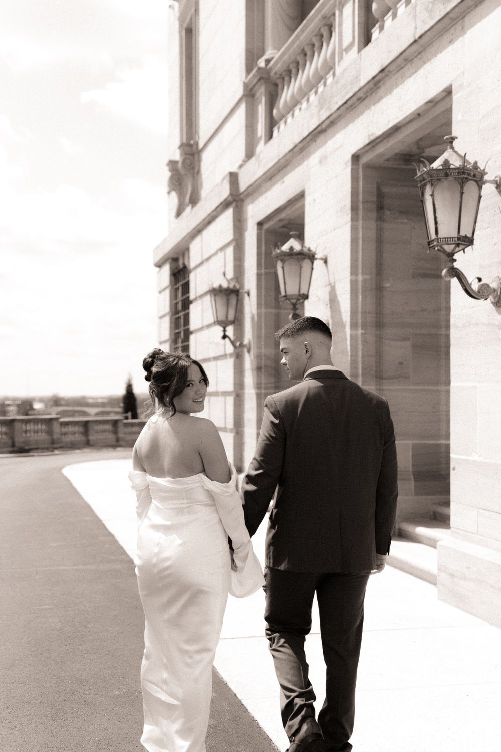 A couple walks hand in hand along an elegant colonnade with ornate lanterns on a bright day.