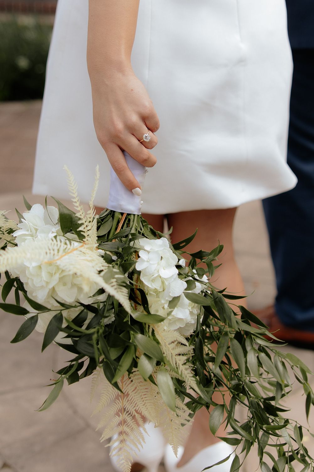 A wedding bouquet with white flowers and cascading greenery held against a white dress.