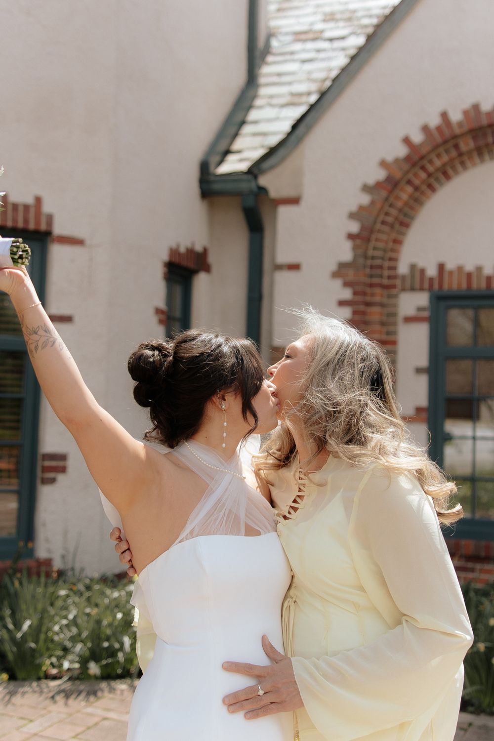 Bride in white dress and companion in yellow share a joyful moment while holding champagne glasses outdoors.
