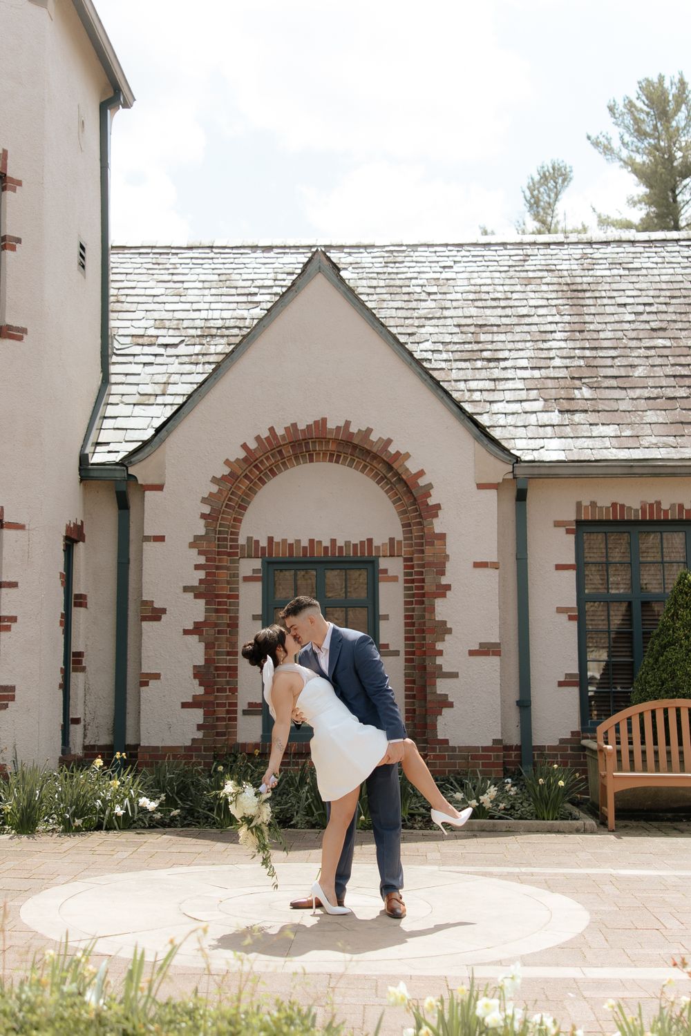 A romantic couple in wedding attire share a kiss while dipping in front of a charming cottage-style building.