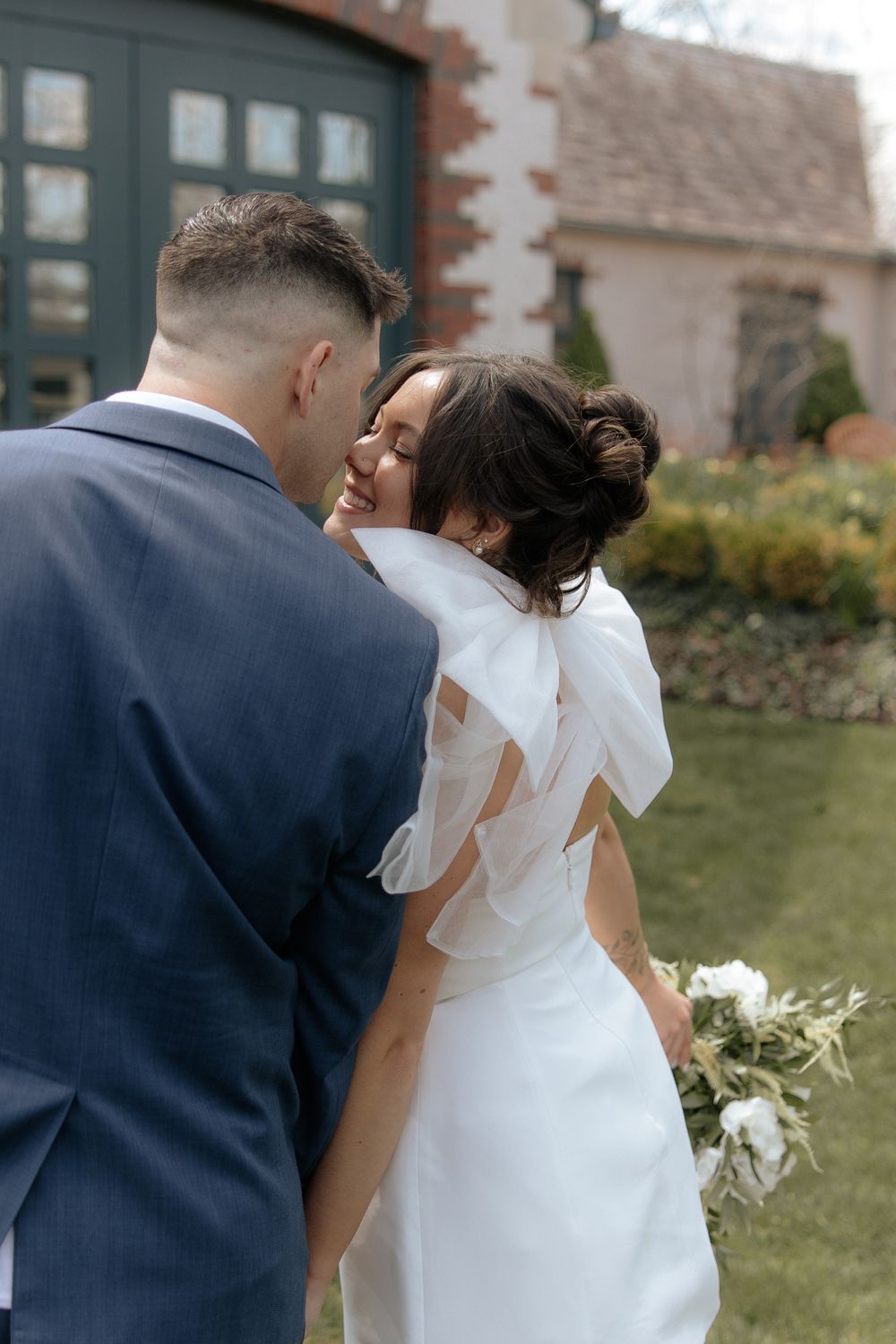 Newlywed couple in navy suit and white dress embracing in a garden setting.