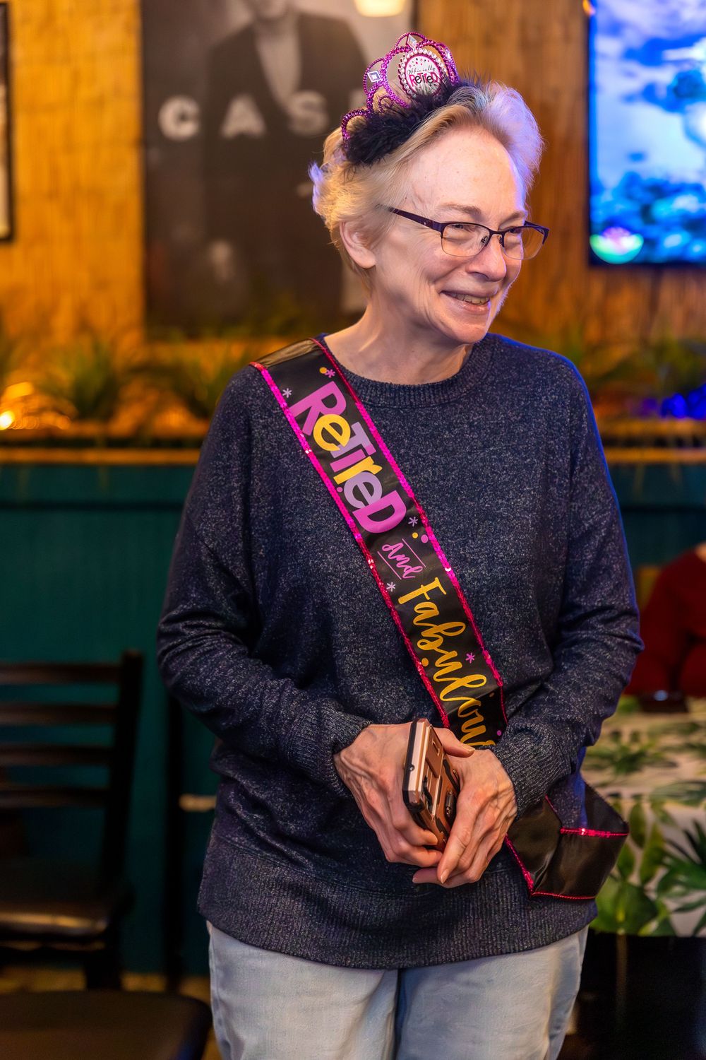 A guest wearing a celebratory sash over a gray sweater smiles warmly at a birthday gathering.