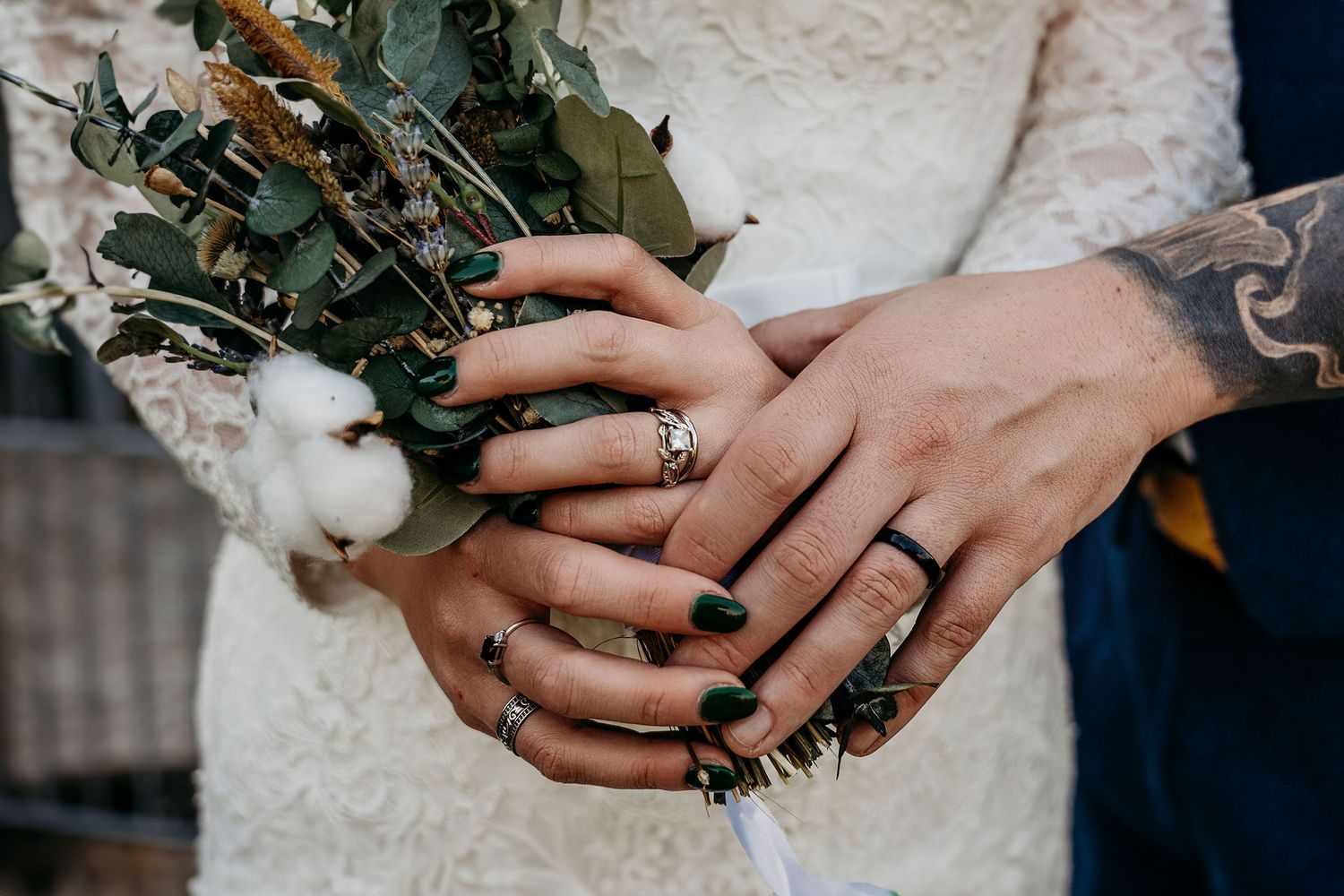 Close-up of hands with wedding rings holding a rustic bouquet with greenery.