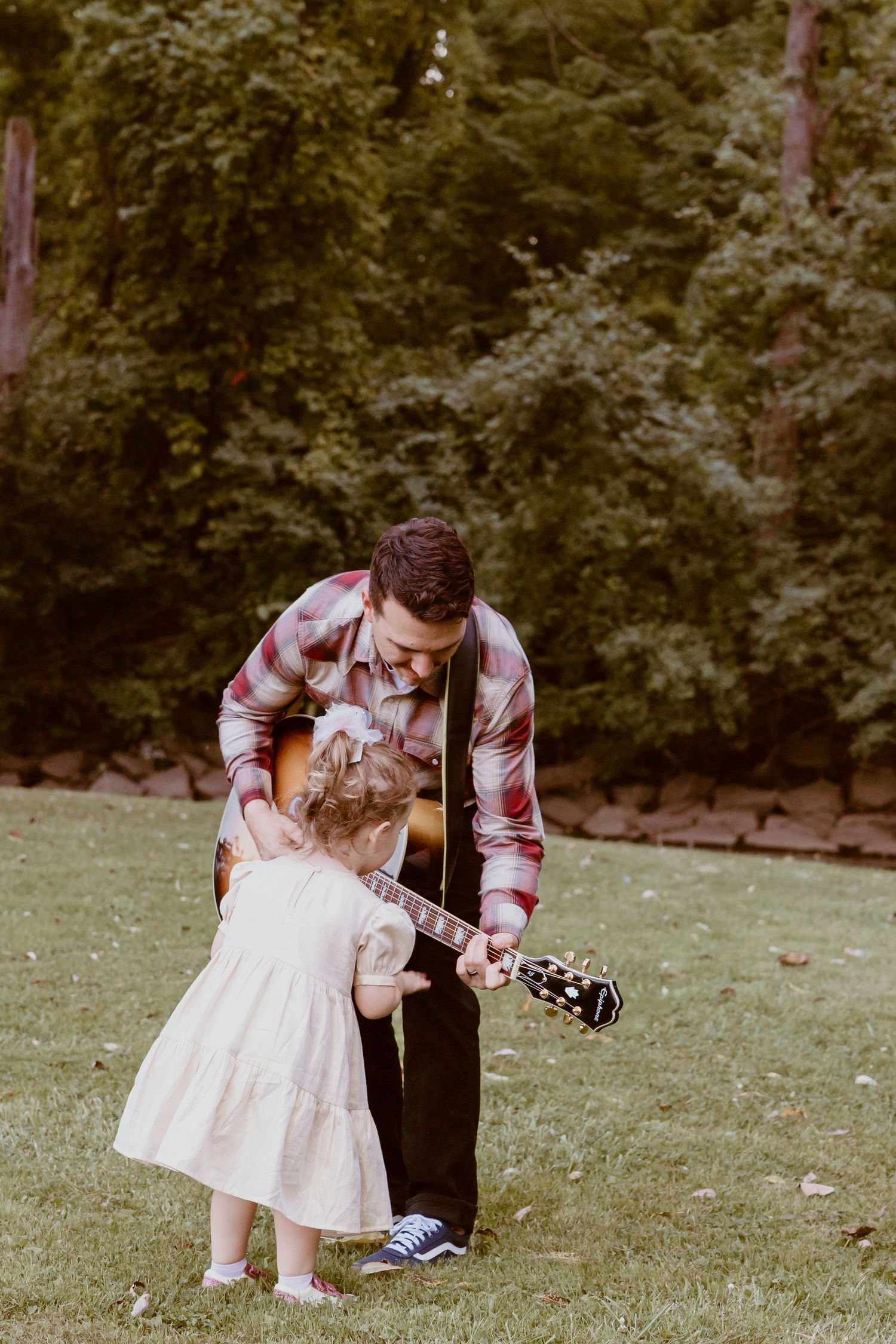 A person in a plaid shirt shows a young child in a white dress how to play guitar on a grassy lawn.