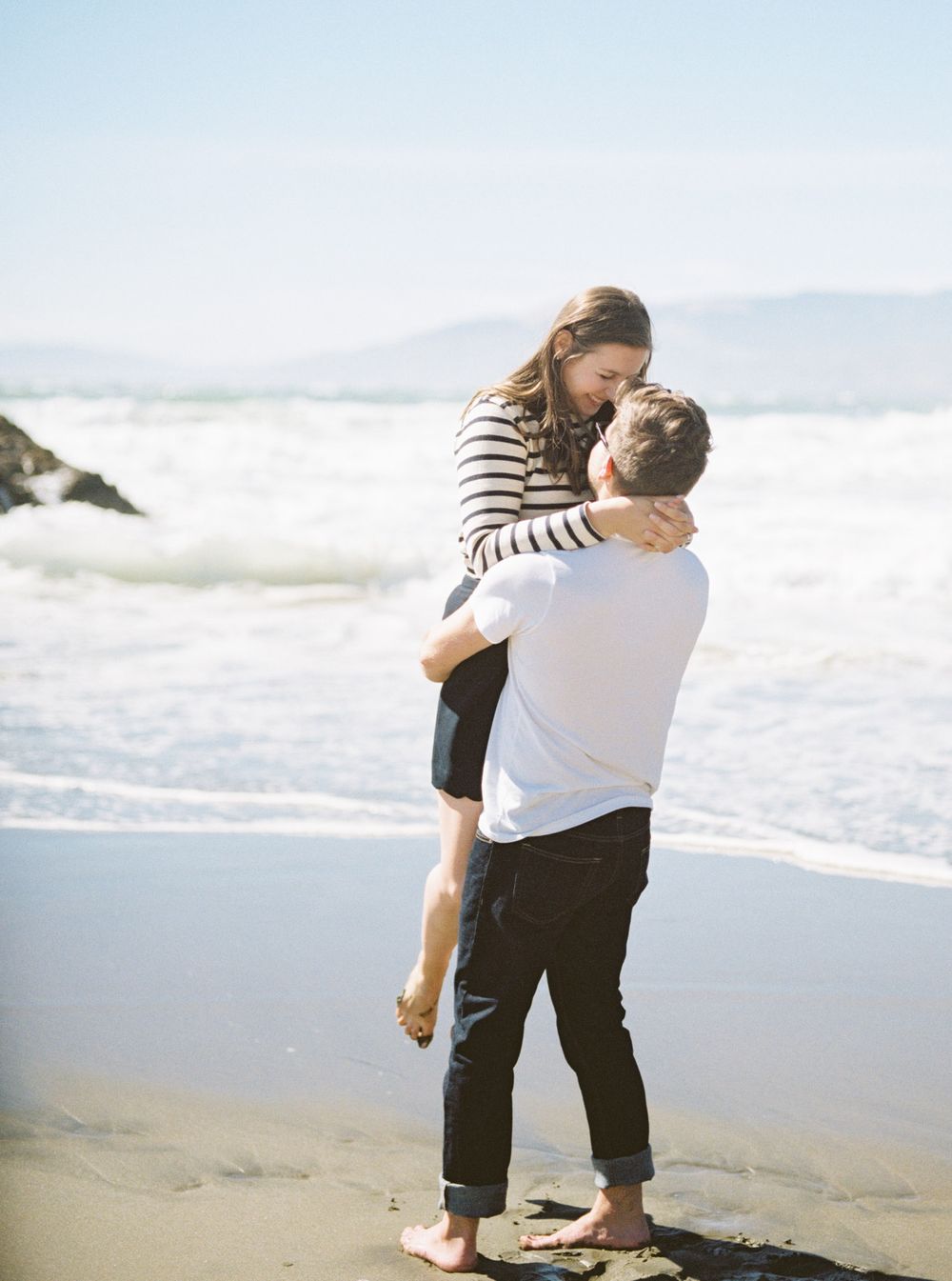 Couple embracing on a beach with crashing waves and ocean in the background.