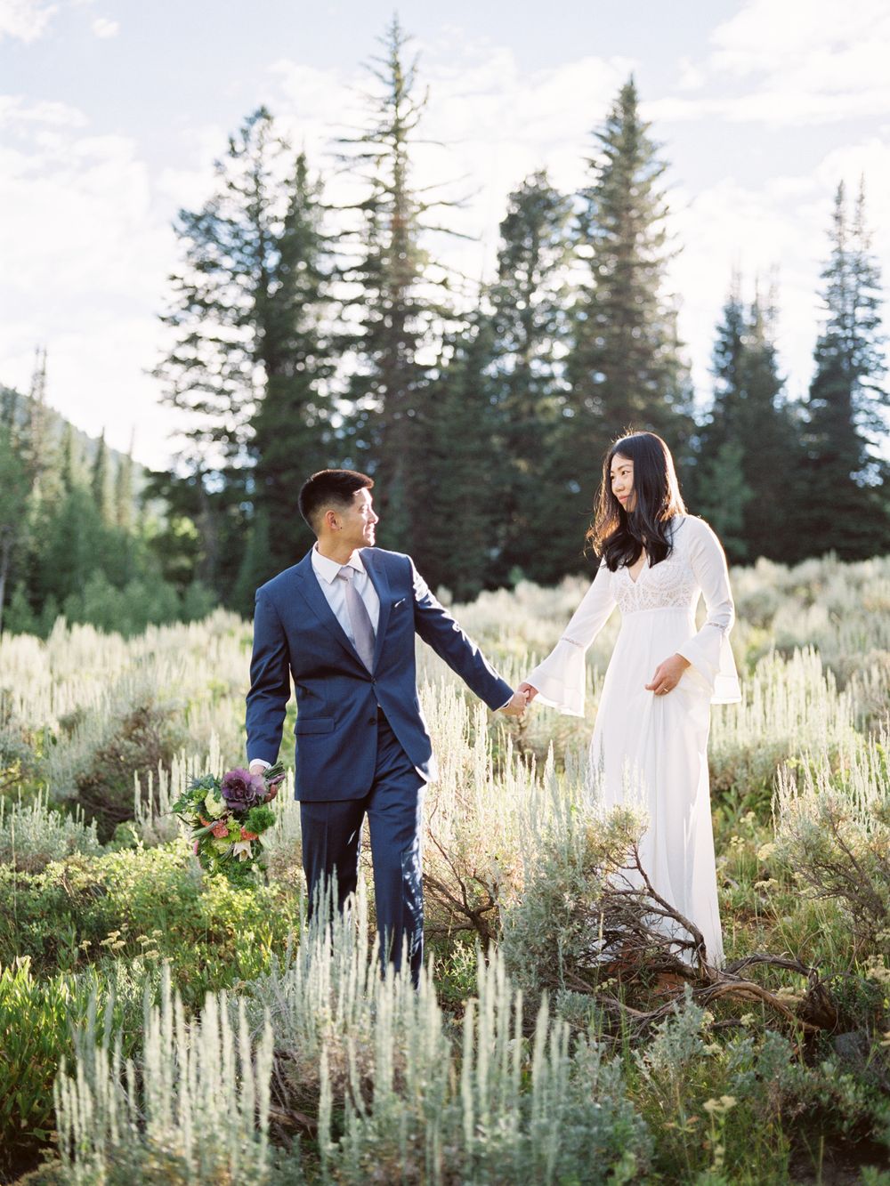 Couple walking hand in hand through a mountain meadow with evergreen trees in the background.