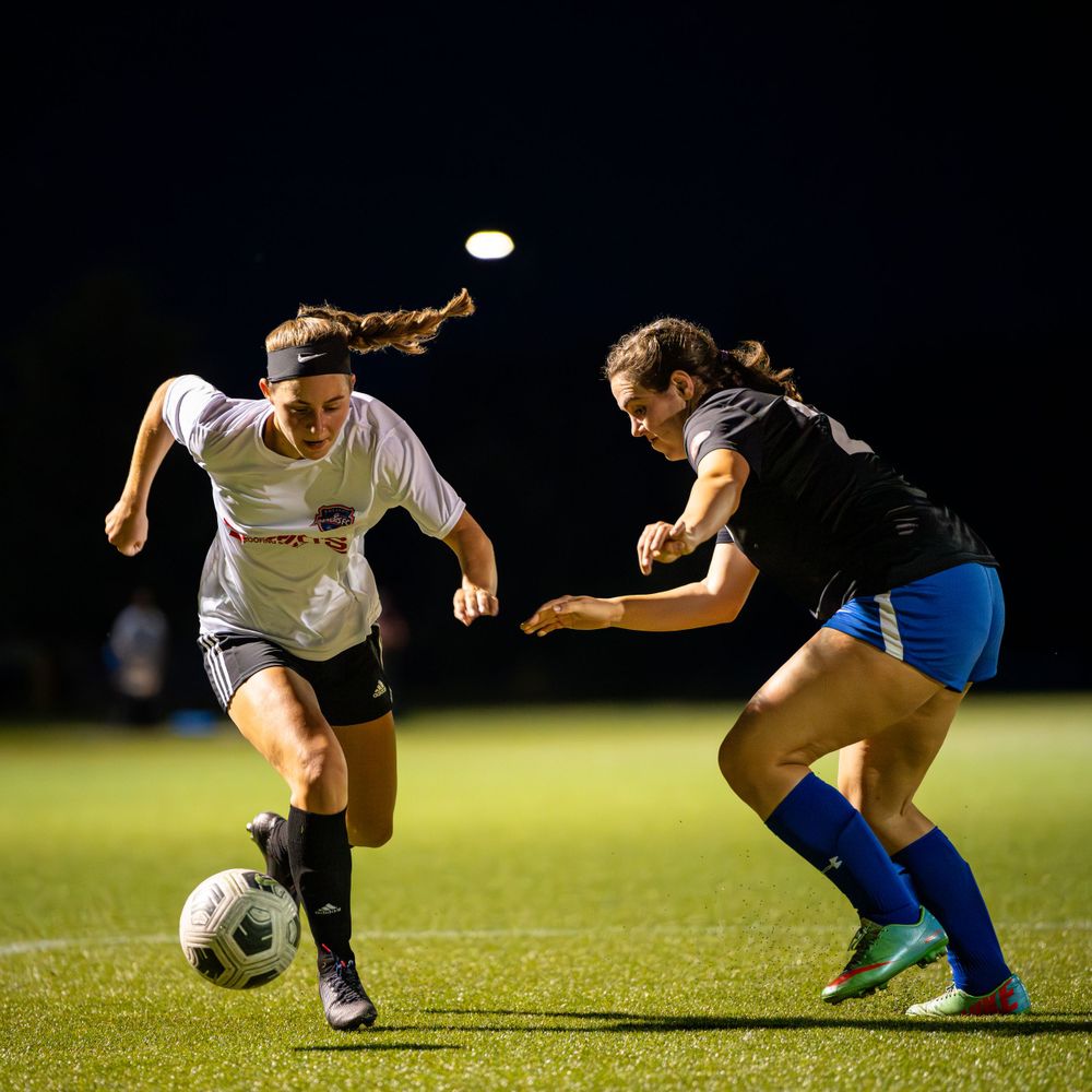 Two players battle for possession of soccer ball during nighttime match on illuminated field.