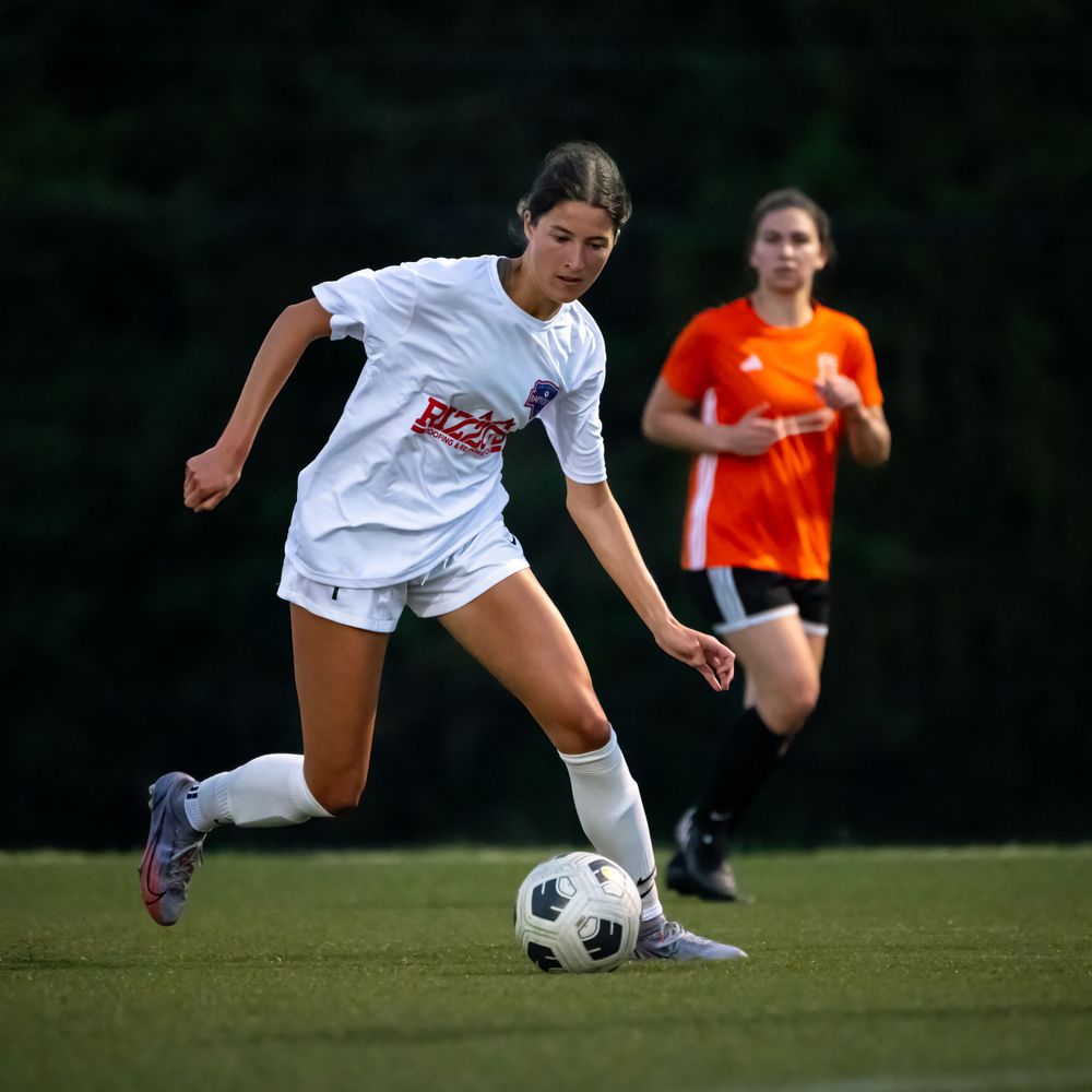 Soccer player in white uniform controls ball while opponent in orange approaches from behind.