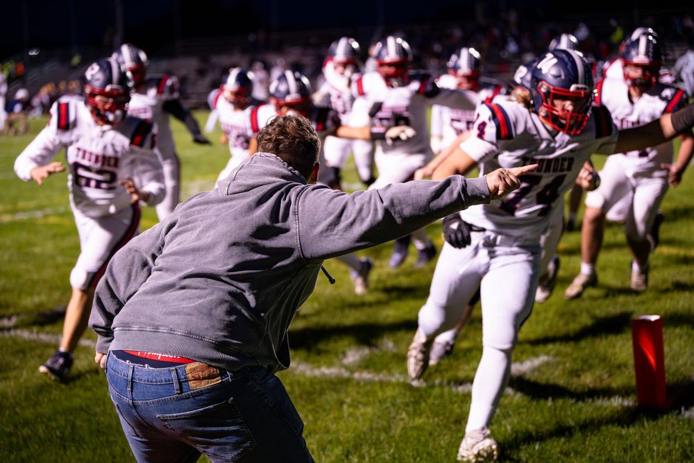 Coach gestures energetically on sideline as football players rush past in white uniforms.