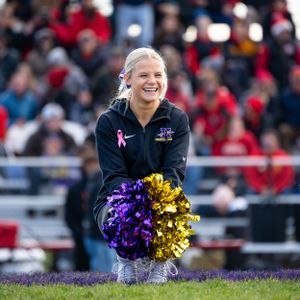 A cheerleader on a sports field holds purple and gold pom poms while wearing a black jacket with a pink ribbon.