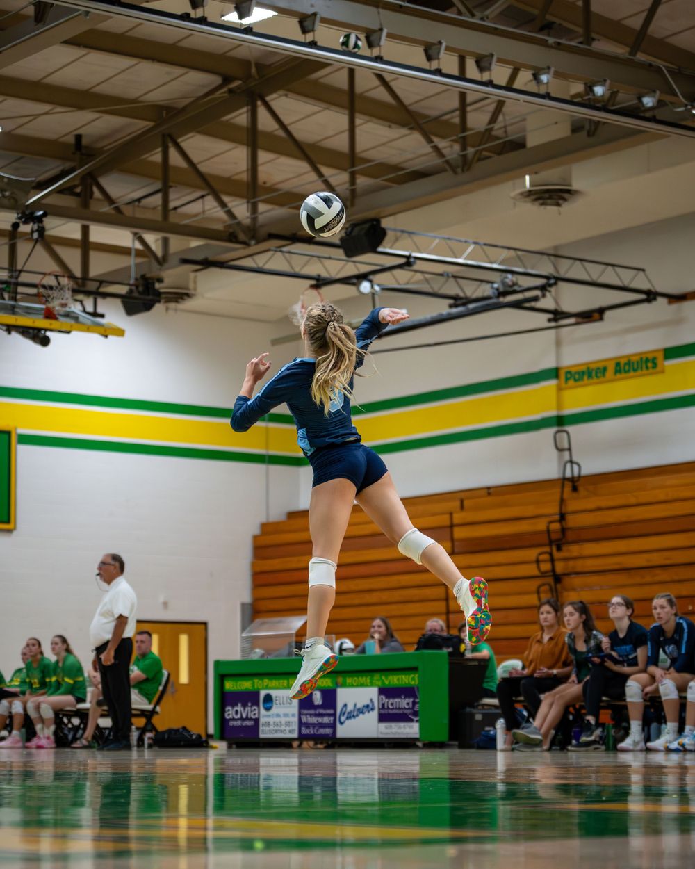 Volleyball player leaps high to spike ball during match in school gymnasium.