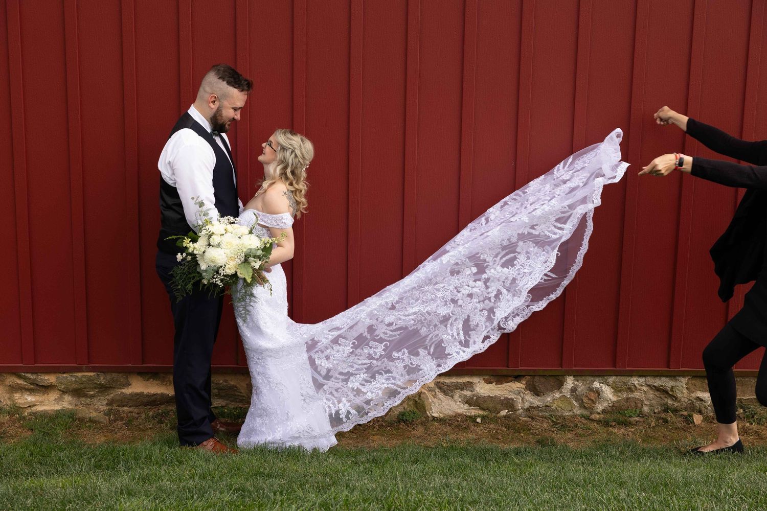 A wedding photographer captures a flowing lace veil against a rustic red barn backdrop as a couple poses.