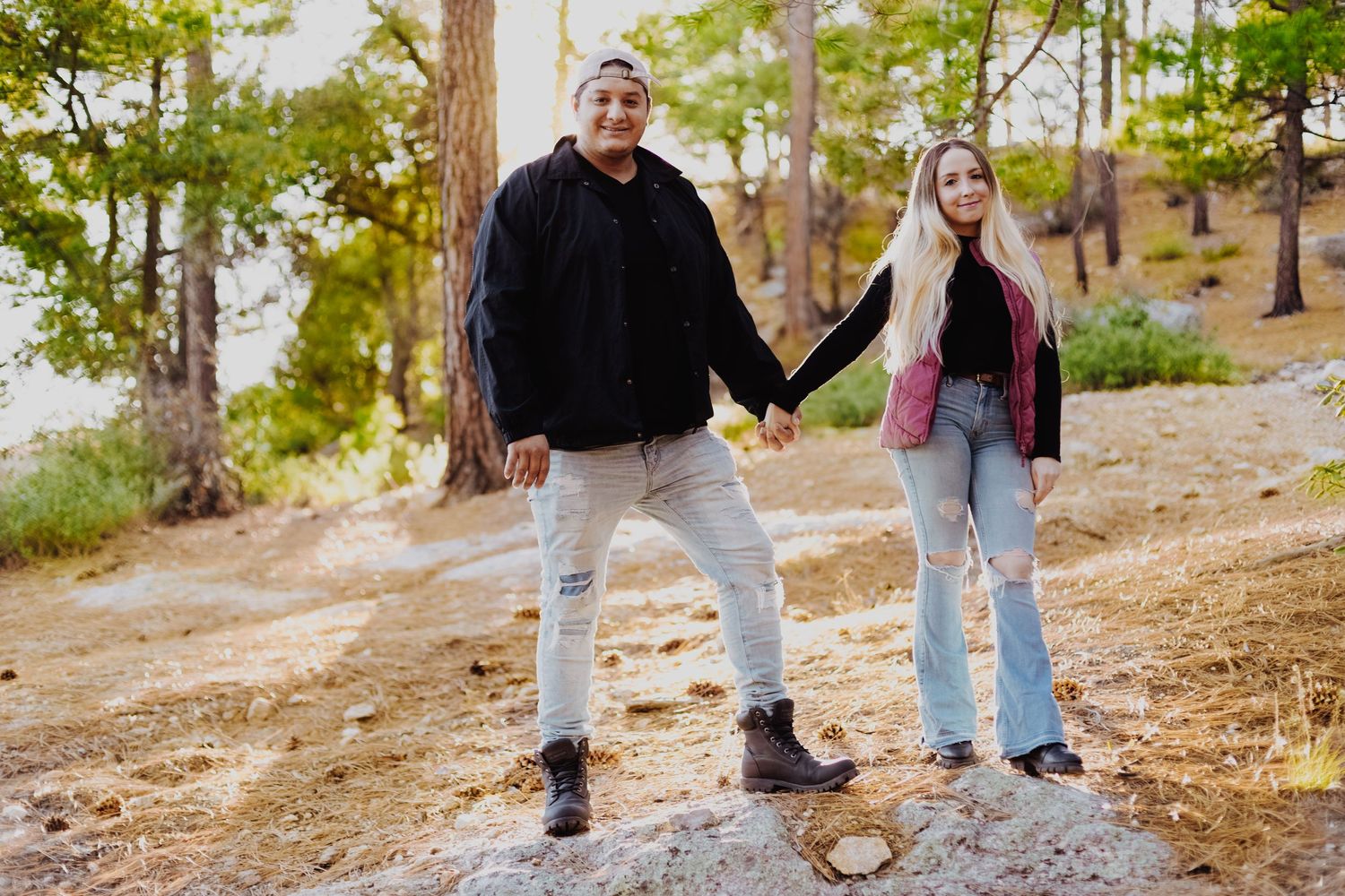 A casual couple holding hands while standing on rocks in a wooded area.
