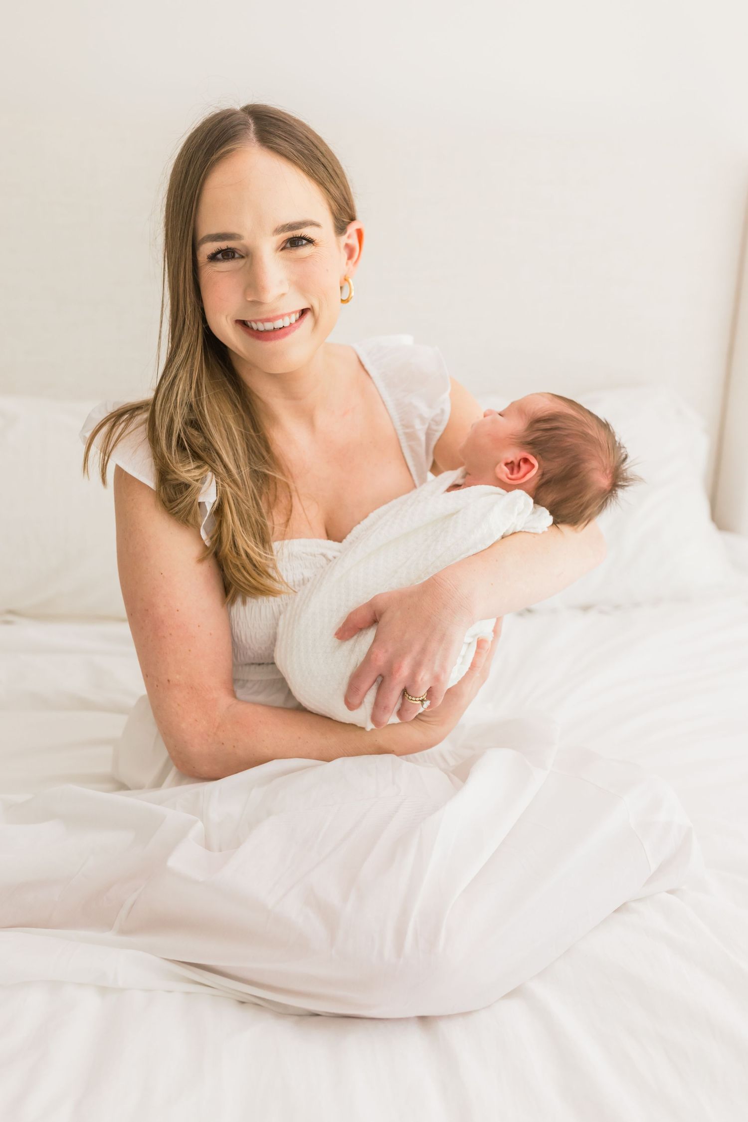 A mother in white dress holds her sleeping newborn while sitting on white bedding with natural light.