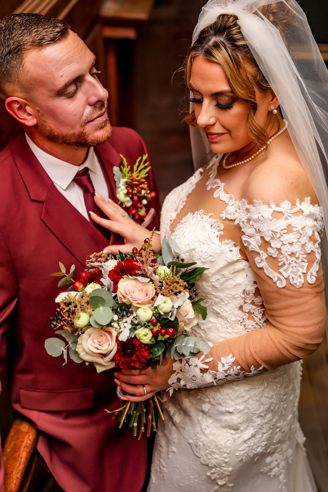 A romantic wedding portrait showing a bride in lace and groom in burgundy suit embracing with a floral bouquet.