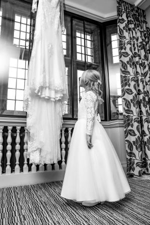 A elegant white lace wedding dress hangs near a window while another is worn in a vintage-style room.