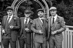 Groomsmen in vintage-style suits and flat caps pose together at an outdoor wedding venue.