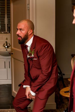 A groom in a burgundy suit jacket poses expressively during a wedding celebration.