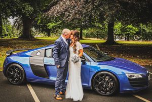 A wedding couple poses with a bright blue Audi R8 sports car in a tree-lined parking area.