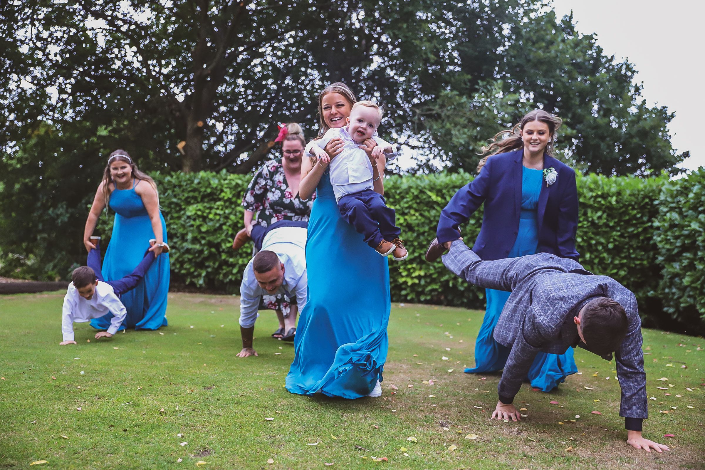 Wedding party members playing and posing on grassy lawn wearing bright blue outfits.