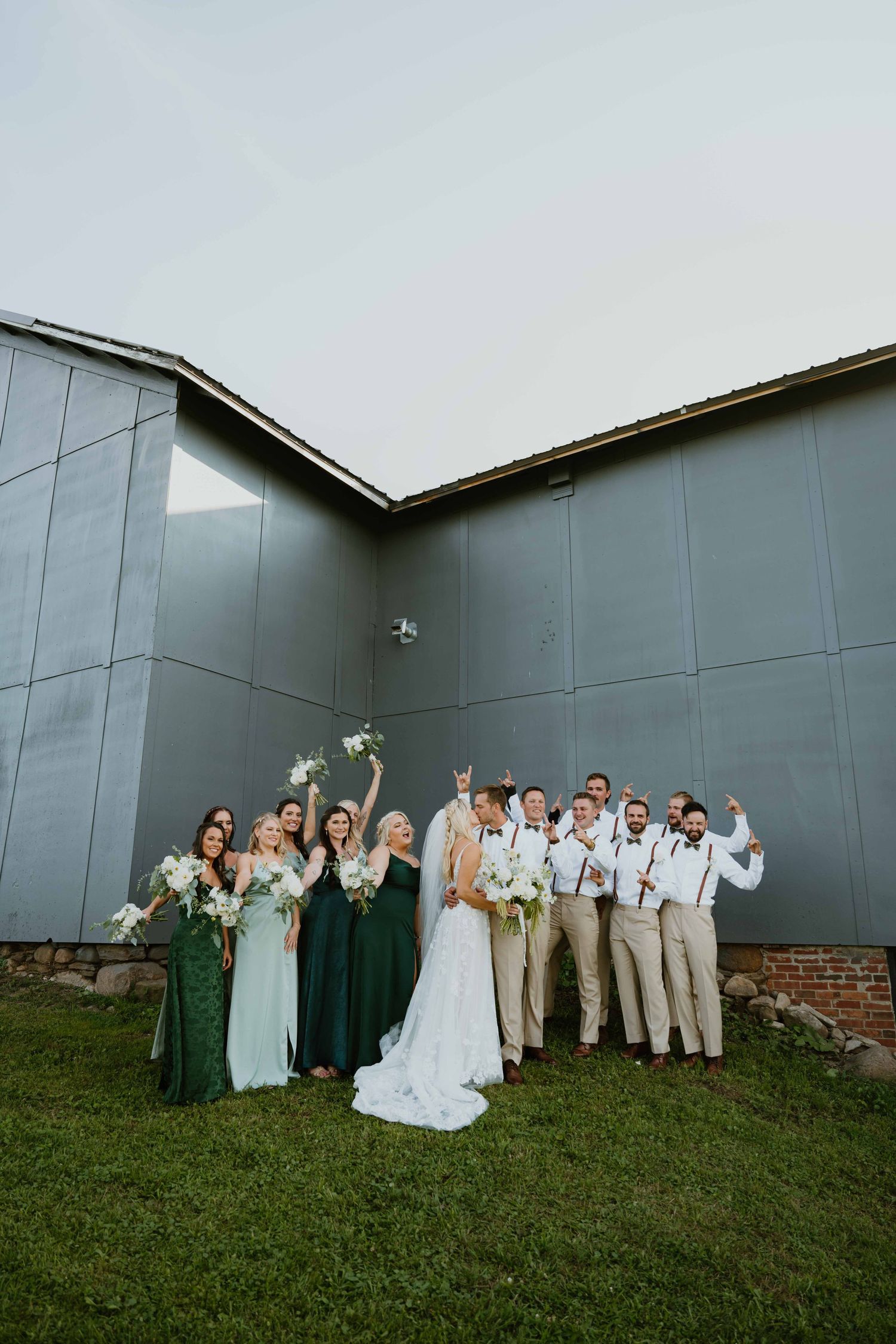 Wedding party celebrating in front of modern gray barn, bridesmaids in green dresses, groomsmen in tan suits.