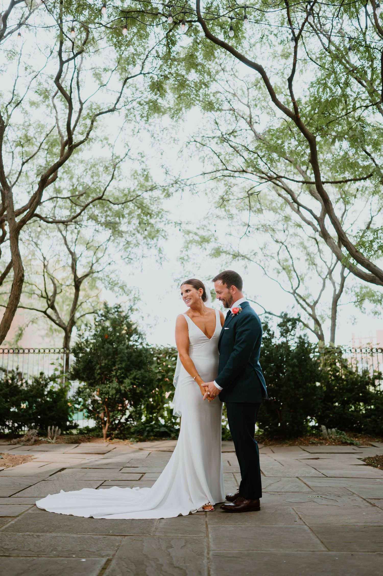 Bride and groom hold hands and gaze at each other beneath lush green trees in a garden wedding portrait.
