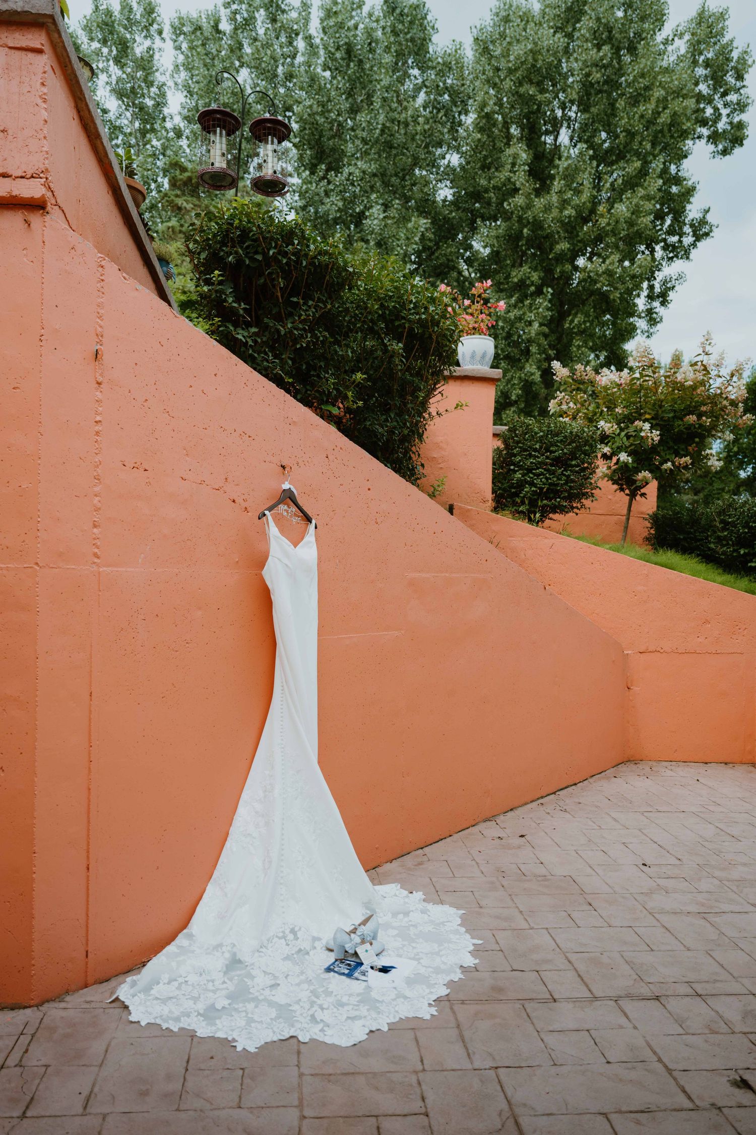 Elegant lace wedding gown hangs against a terracotta wall with a long flowing train.